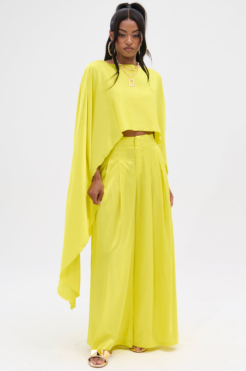 Dressed in a vibrant yellow glossy satin ensemble, featuring the COCKTAIL HOUR BLOUSE with a high-low hem and wide-legged pants, an individual stands confidently. They accessorize with layered gold necklaces and open-toed sandals, set against a plain white background.