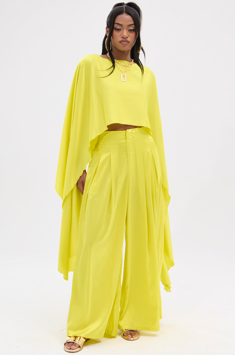 A person is styled in a bright yellow COCKTAIL HOUR BLOUSE with wide-legged pants and a cape-like top featuring a flowy, high-low hem. Their long dark hair complements the gold jewelry—necklaces and earrings—and the plain, light backdrop highlights their vibrant ensemble.