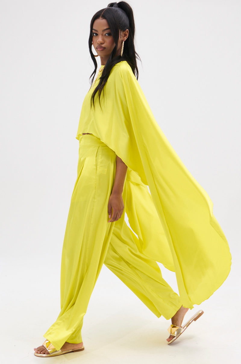 Wearing the COCKTAIL HOUR BLOUSE in bright yellow satin with a high-low hem and cape feature, the person stands in profile. It complements their long dark hair and sandals against a plain light backdrop.