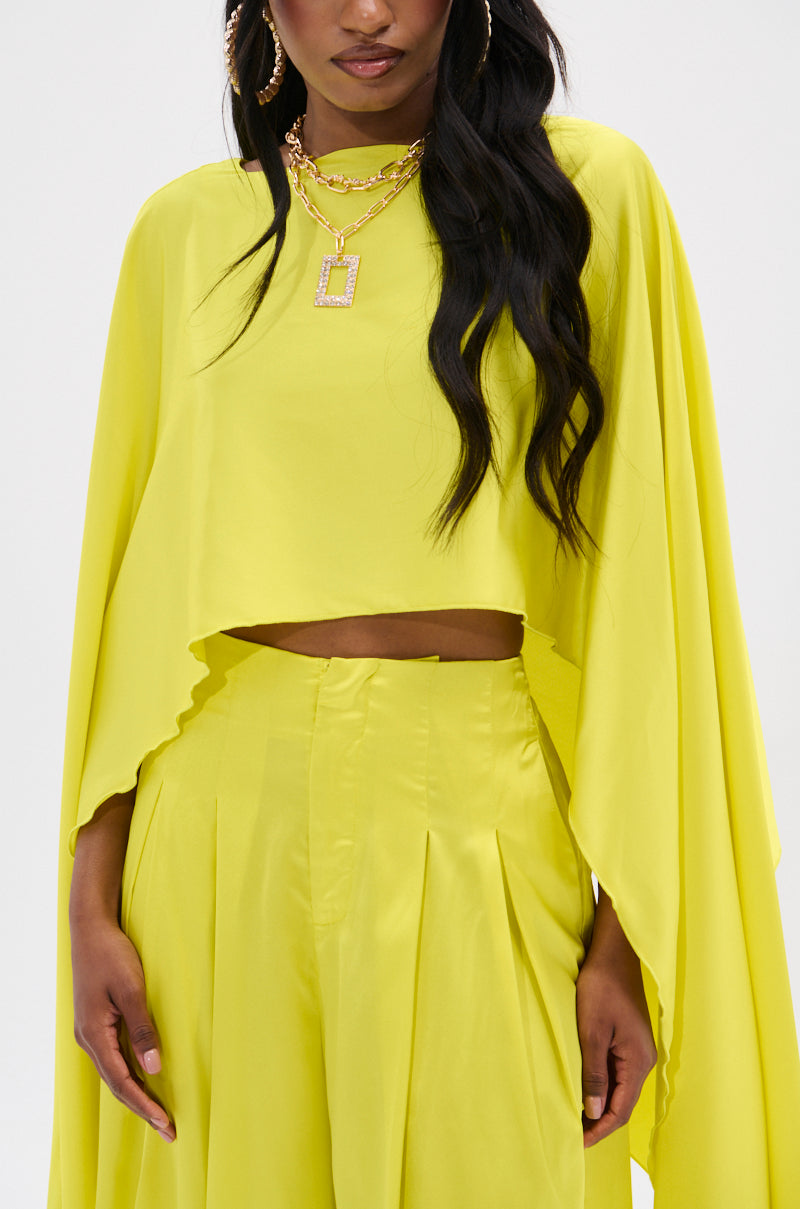 A person models a bright yellow outfit with the Cocktail Hour Blouse, featuring a high-low hem and pleated pants. The loose-fitting cropped top is paired with large hoop earrings and layered gold necklaces against a plain white background that enhances the outfit's vivid allure.