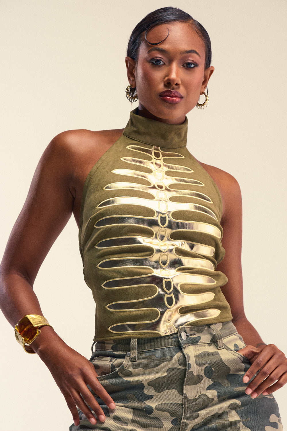 A woman poses confidently in the REMEMBER ME SUEDE TOP, a high-neck olive green suede piece featuring metallic faux leather ribcage detailing. She pairs it with camo pants, hoop earrings, and a gold bracelet, her hair styled sleekly.