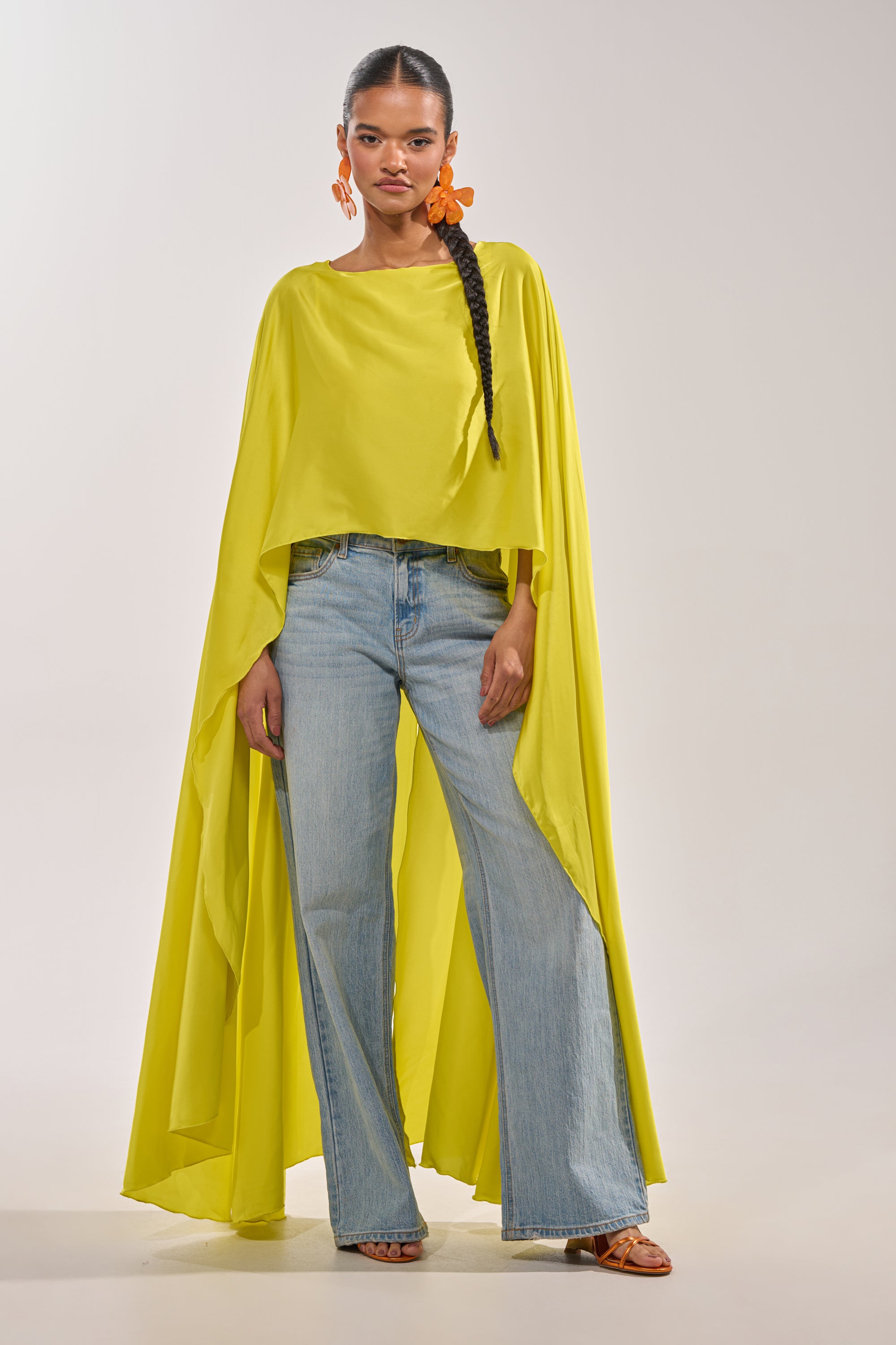 A woman in the SO CHIC CAPE BLOUSE IN YELLOW, light blue wide-leg jeans, orange flower-shaped earrings, and sandals stands confidently against a plain light background. 0
