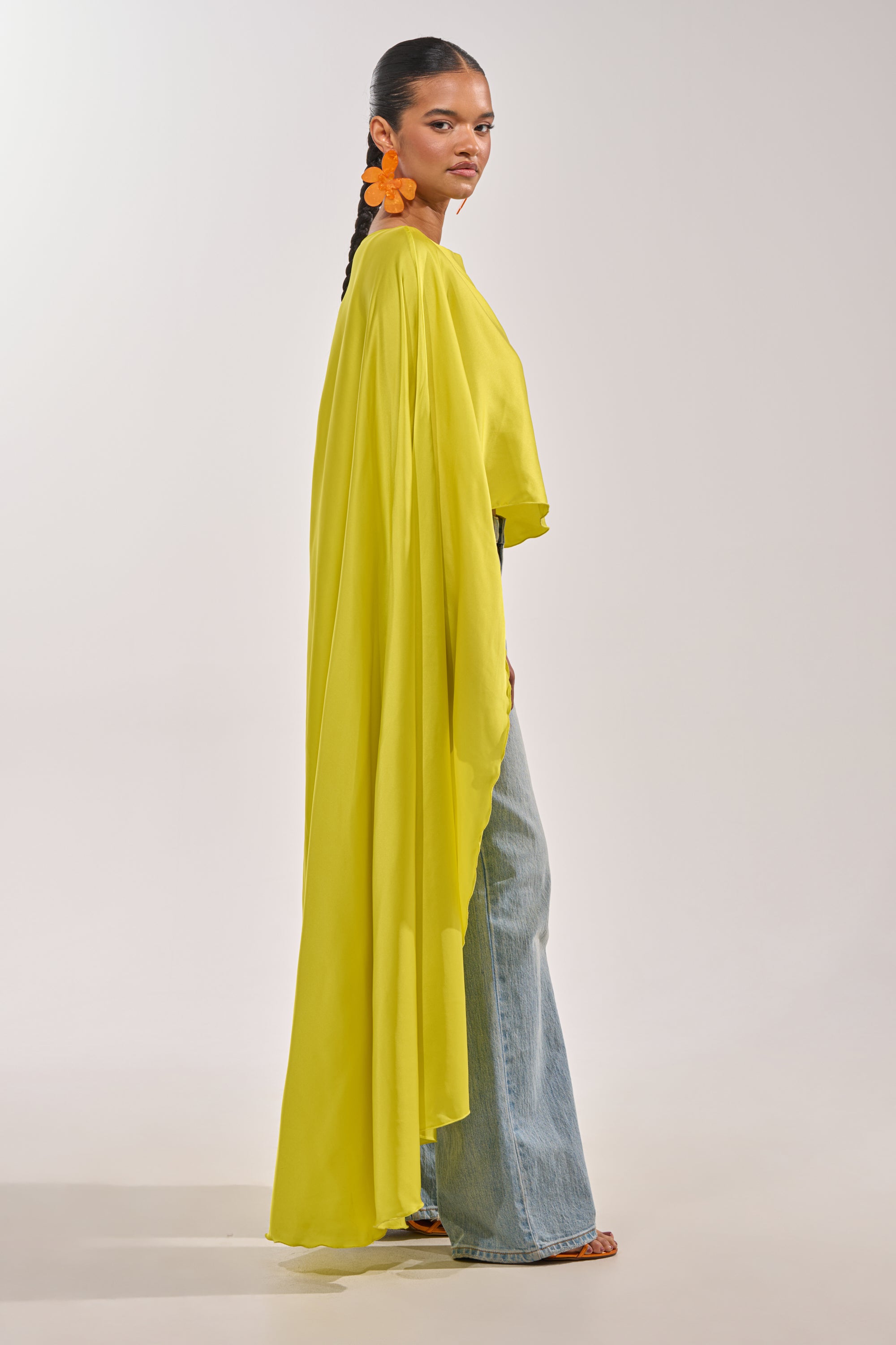 A woman stands sideways in the SO CHIC CAPE BLOUSE IN YELLOW over light blue wide-leg jeans, with sleek hair and large orange flower earrings, set against a plain light background. 1
