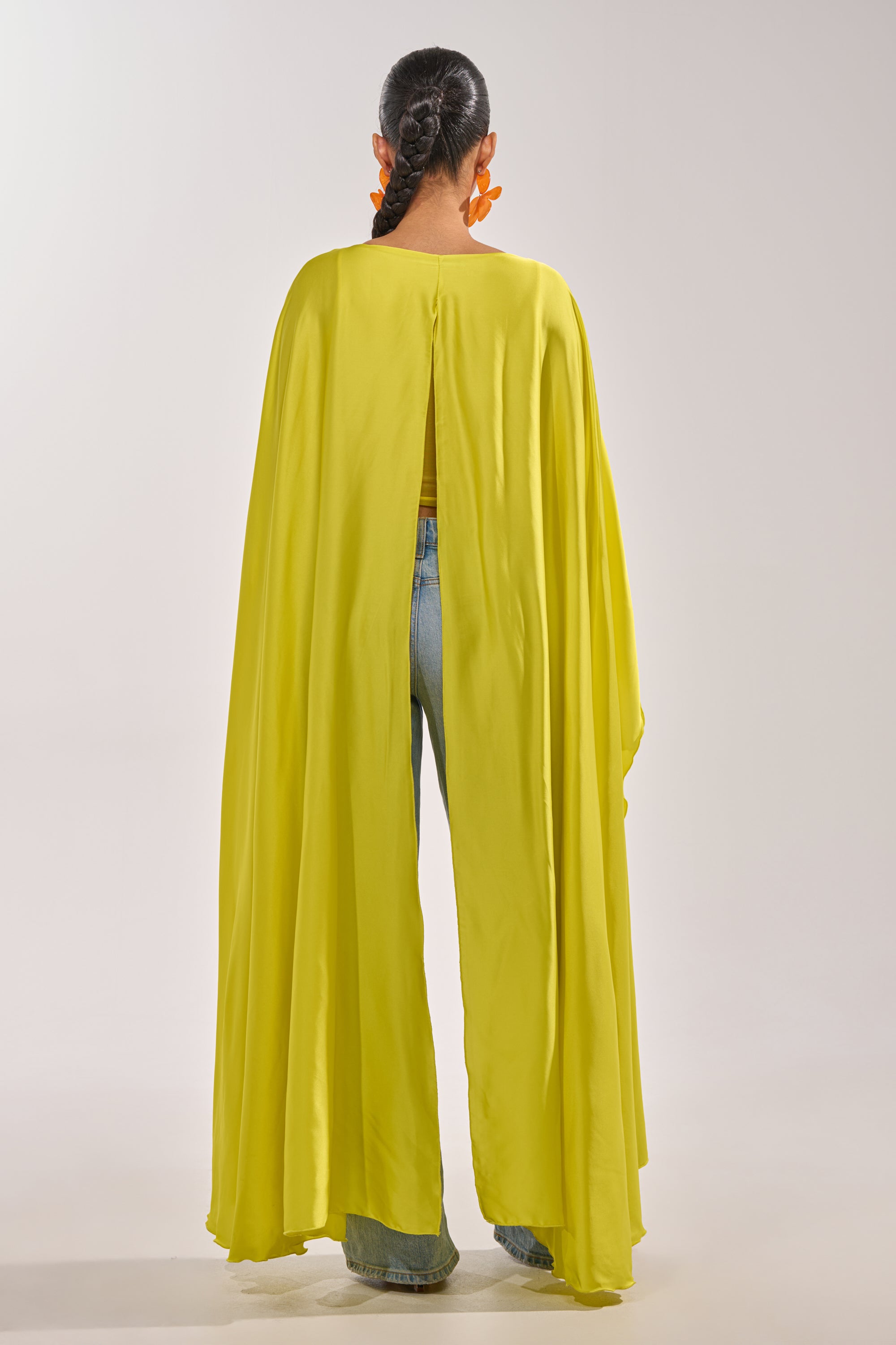 A person with braided hair and orange earrings wears the SO CHIC CAPE BLOUSE IN YELLOW, featuring a draped cape style with a back opening, paired with blue jeans, standing against a plain light background, facing away. 2