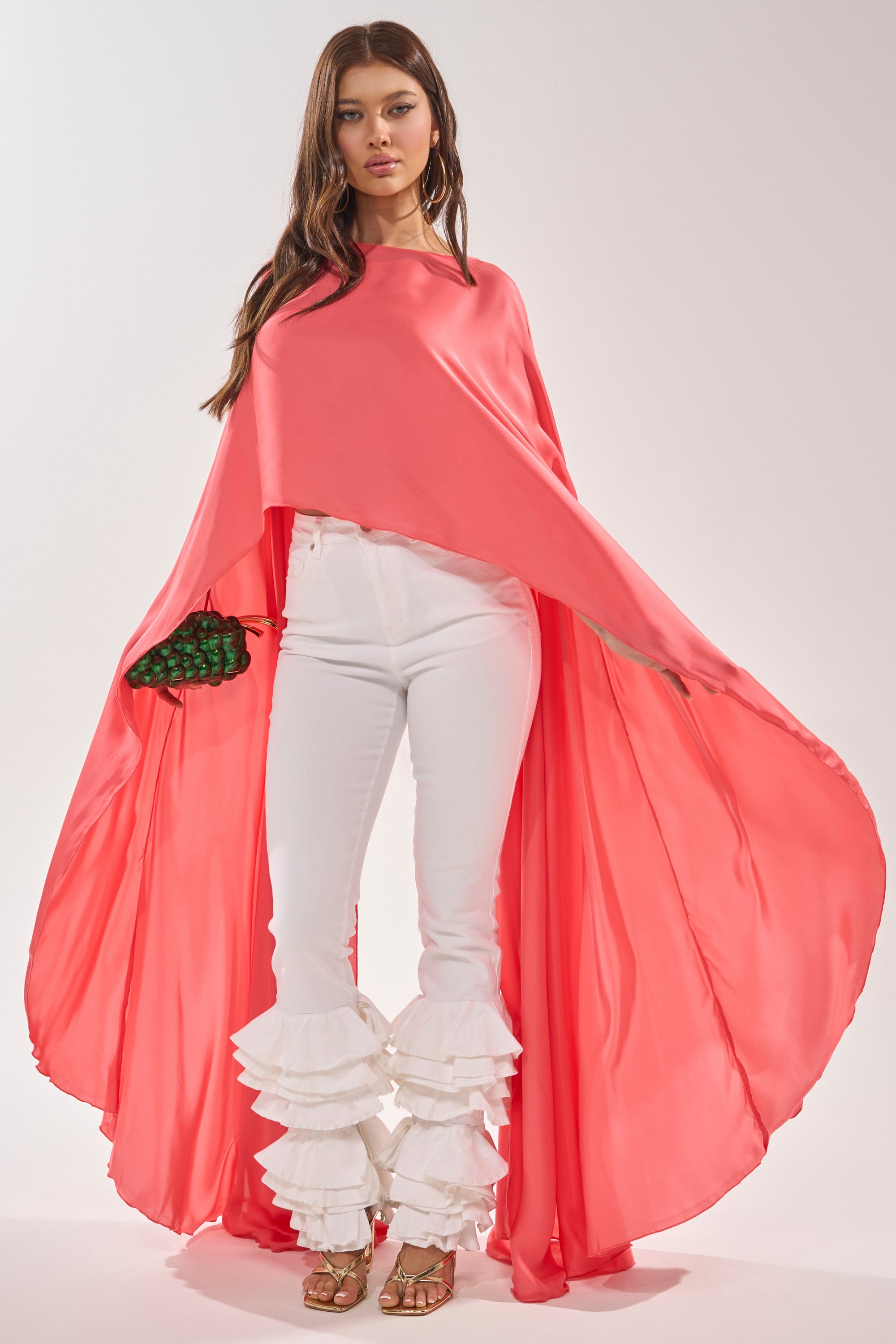 A woman stands confidently in the SO CHIC CAPE BLOUSE, paired with white ruffled pants and gold heels, holding a small green textured clutch against a plain, light background. 0