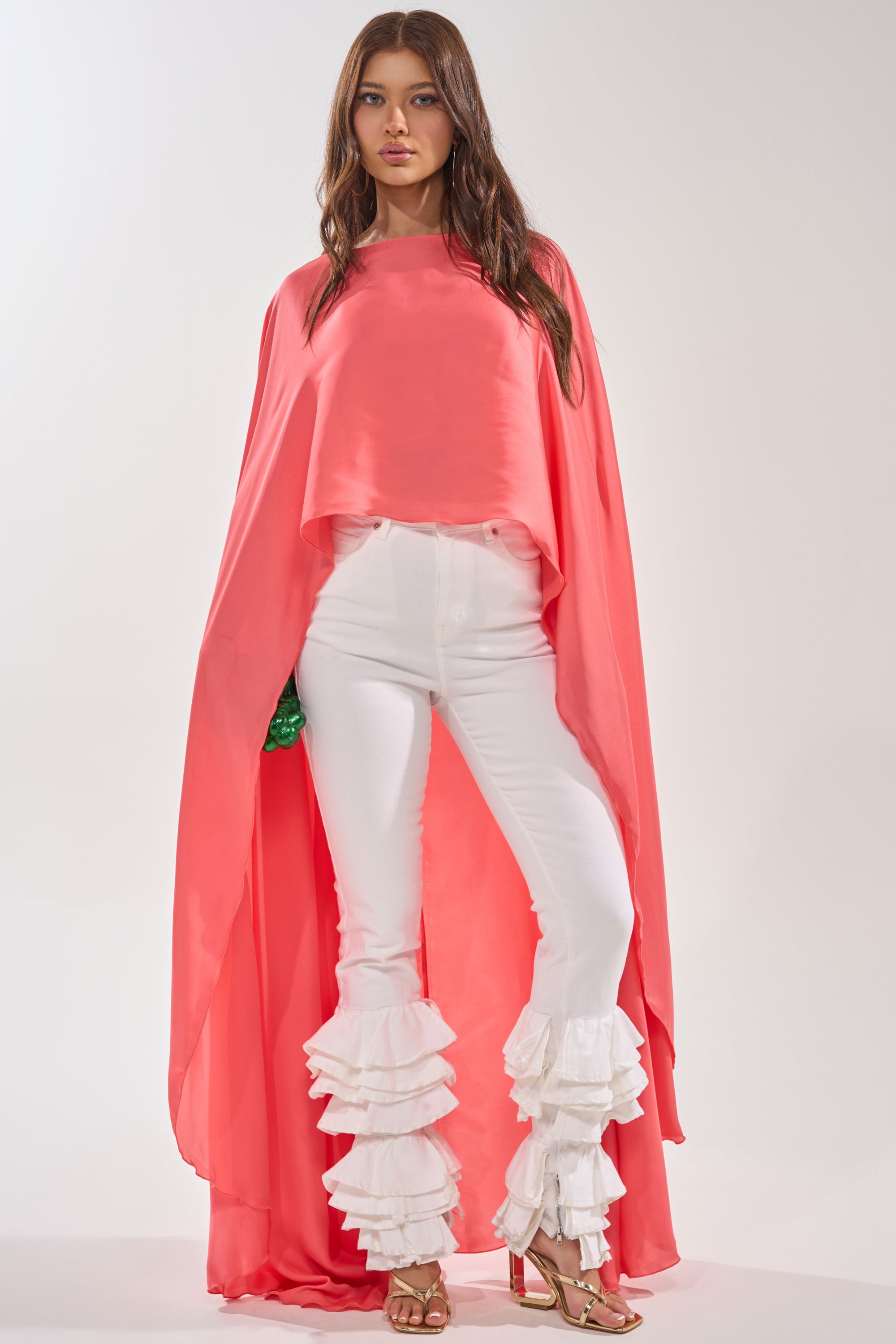 A woman stands against a plain light background wearing the SO CHIC CAPE BLOUSE in coral pink, paired with white pants featuring layered ruffles at the knees and gold high-heeled sandals. 1