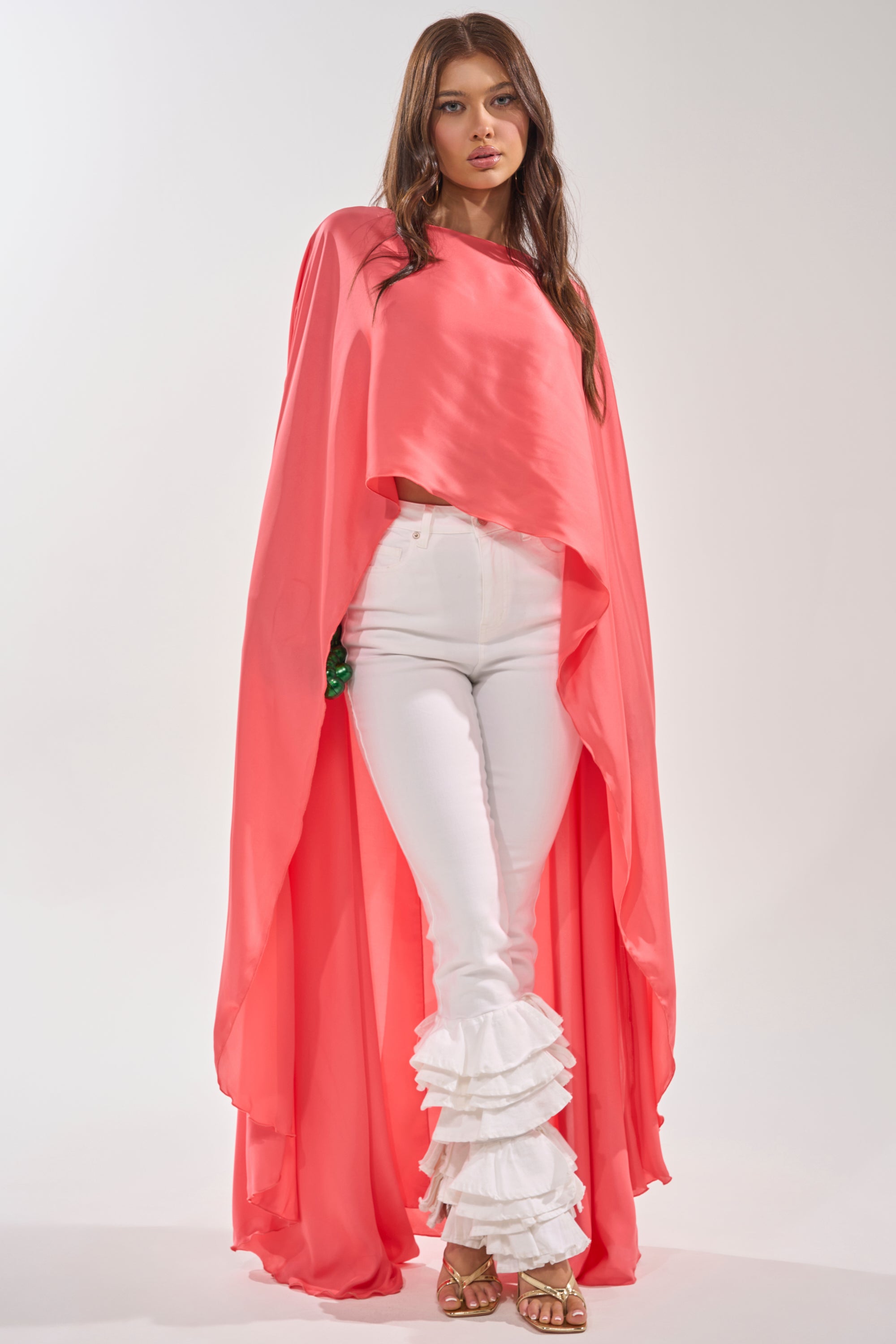 A woman poses in the SO CHIC CAPE BLOUSE, a flowing coral high-low top, paired with fitted white pants featuring layered ruffle ankles and gold sandals, set against a plain light background. 2