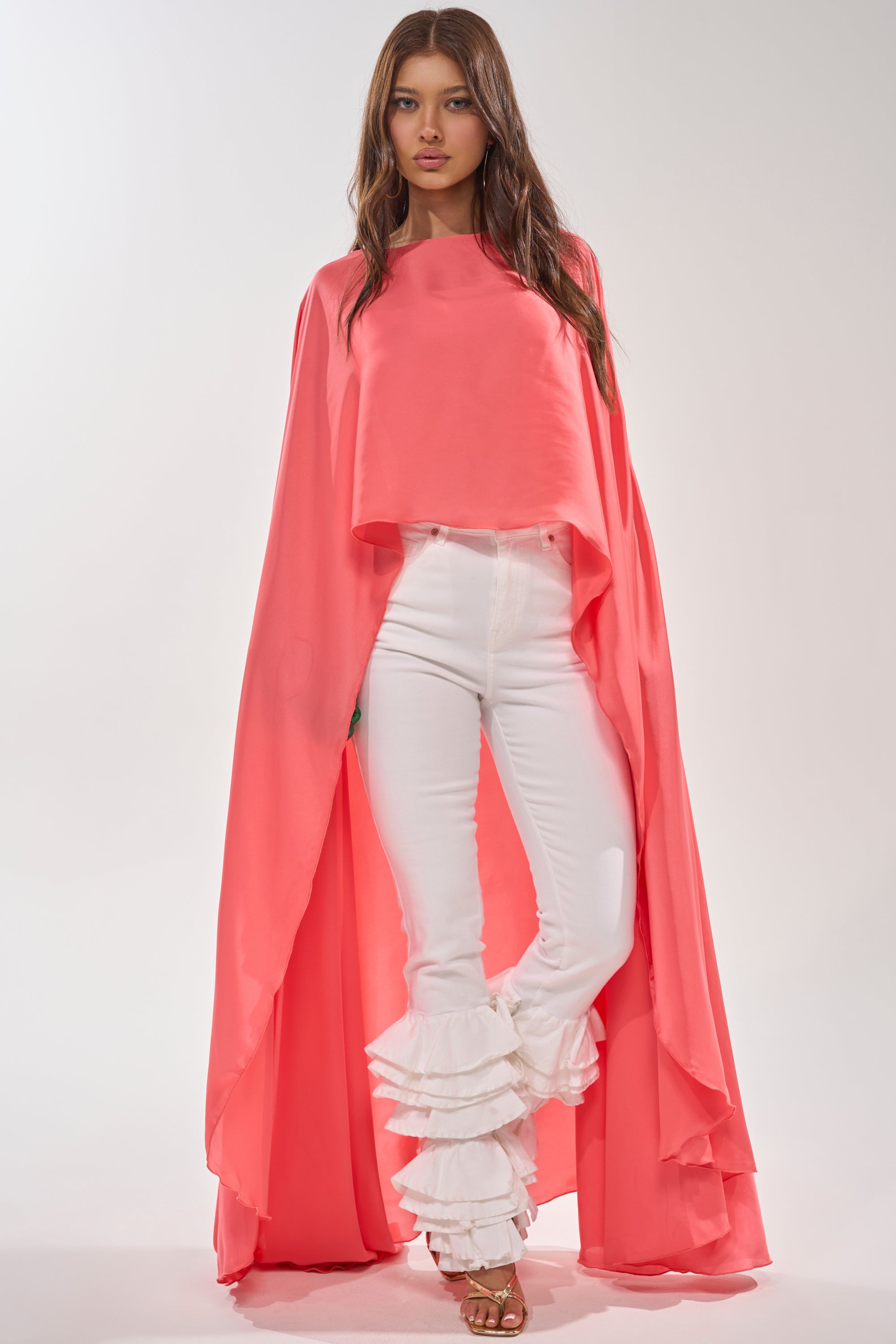 A woman stands against a plain background wearing the SO CHIC CAPE BLOUSE in coral pink, styled with white high-waisted pants featuring layered ruffle ankle details and open-toed heels. 3