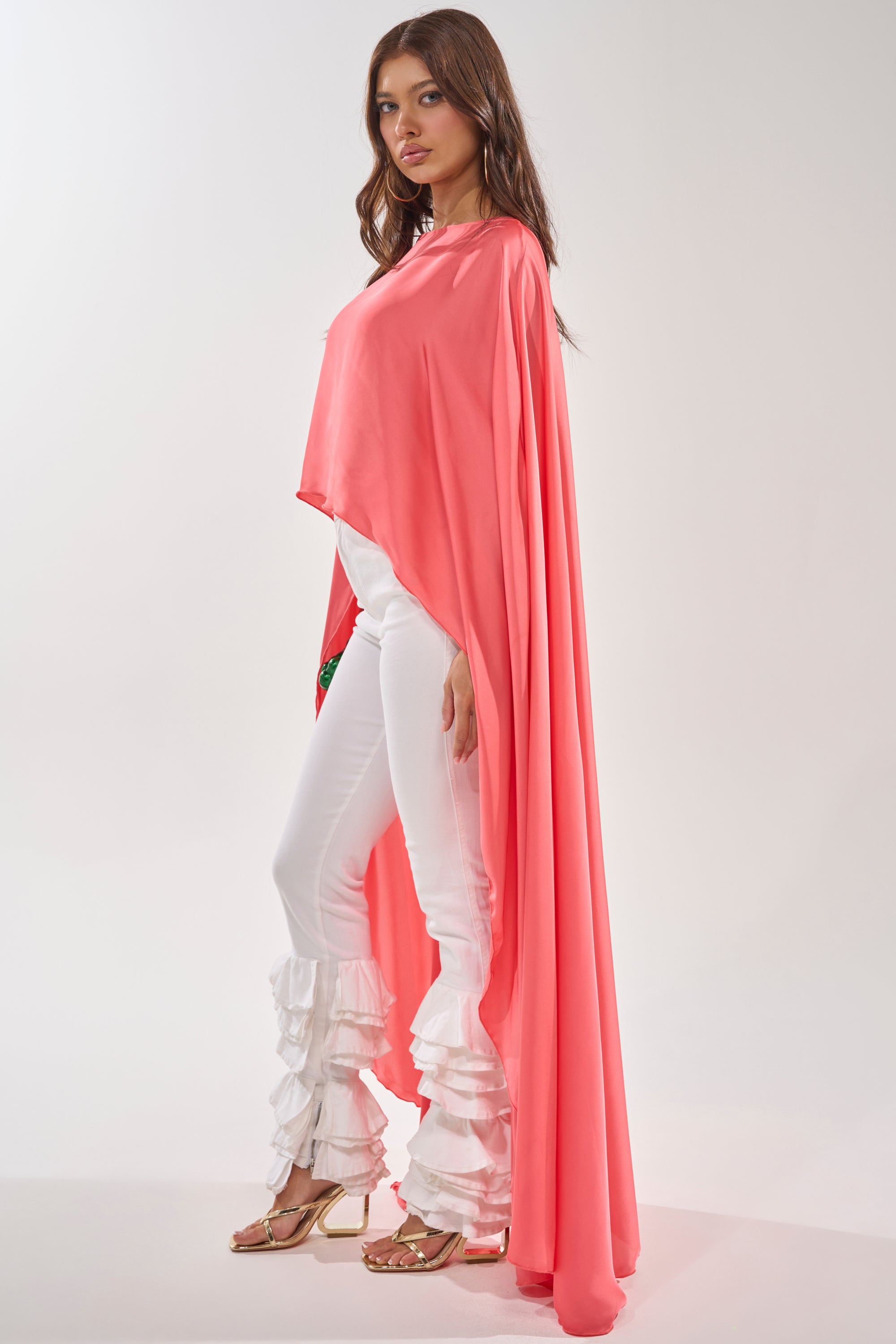 A woman in profile models the SO CHIC CAPE BLOUSE in coral over a white outfit with ruffled, layered flared pants and gold sandals, set against a plain light background. 4