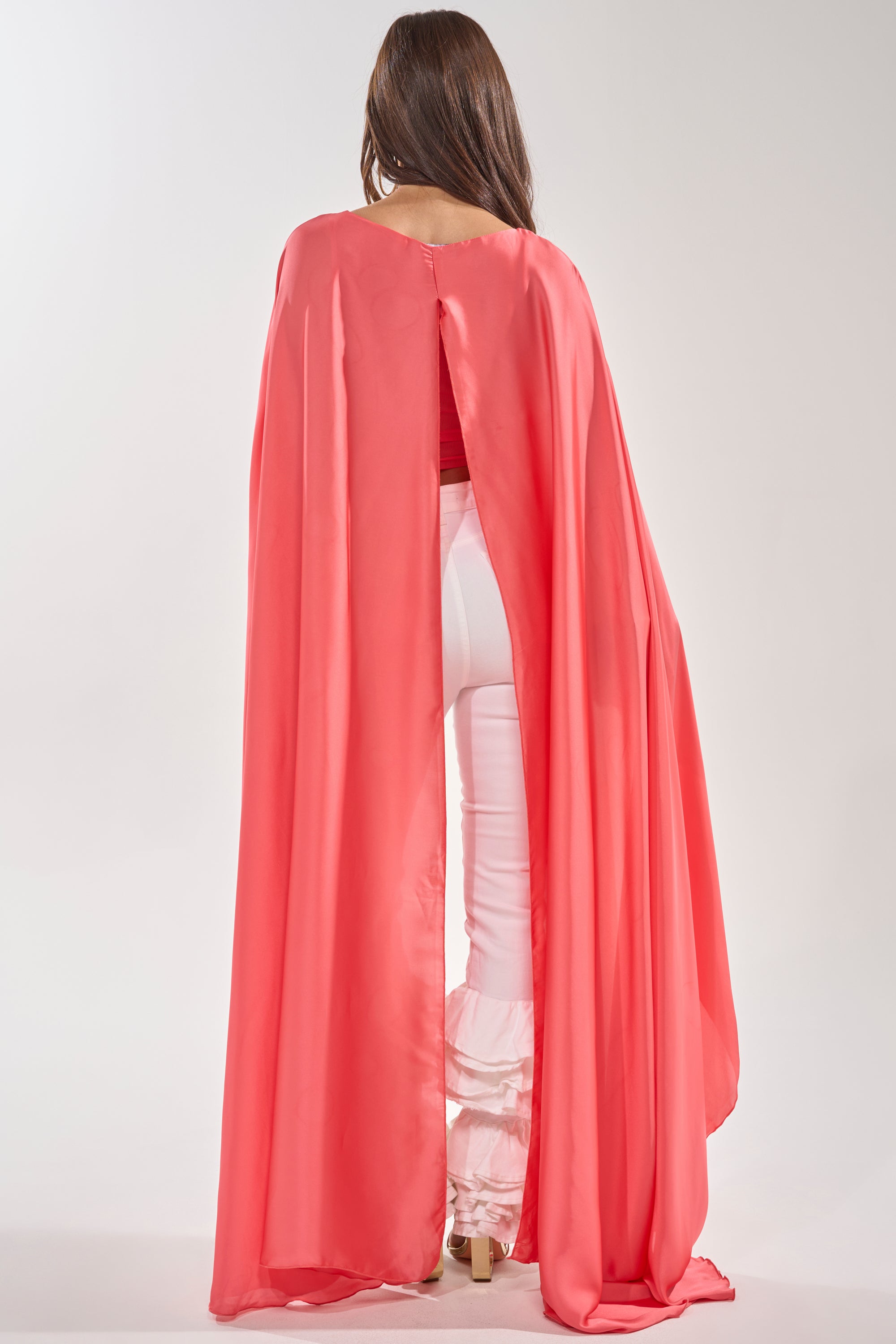 A woman with long brown hair stands against a plain light background, wearing the SO CHIC CAPE BLOUSE in coral over a red top and white ruffled-bottom fitted pants. She is facing away from the camera. 5