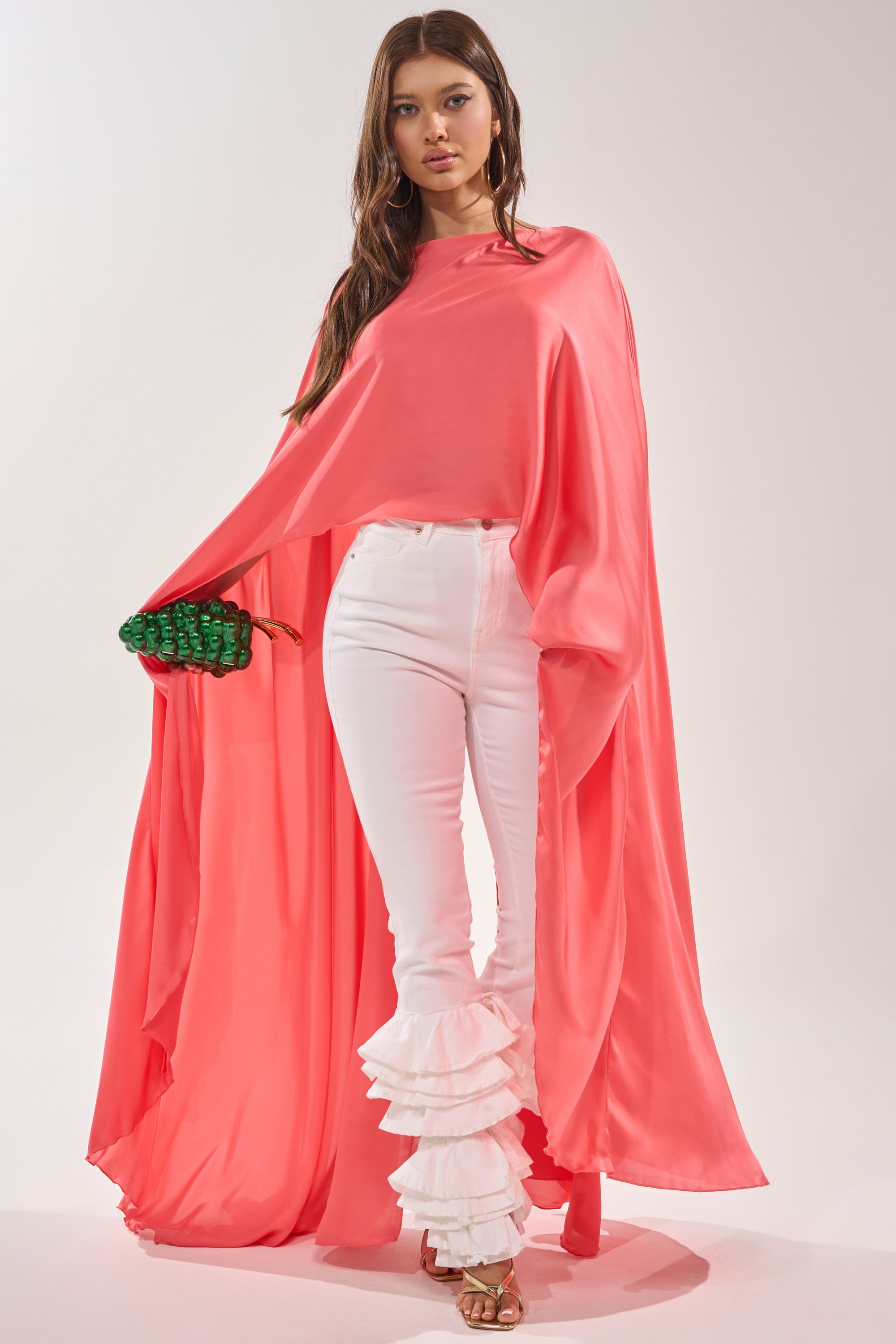 A woman wears the SO CHIC CAPE BLOUSE in vibrant coral, paired with fitted white ruffle-hem pants, gold sandals, and holds a small green textured clutch against a plain background. 6