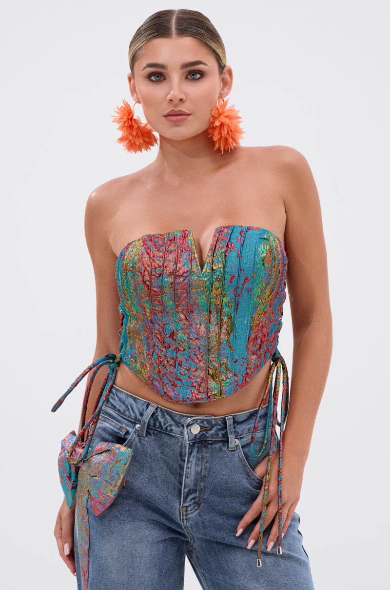 A woman wears the PRETTY GIRL PROBLEMS CORSET, featuring a colorful party style, strapless sweetheart neckline, and lace-up sides, paired with blue jeans. She completes the look with slicked-back hair and large orange flower earrings against a plain light background.