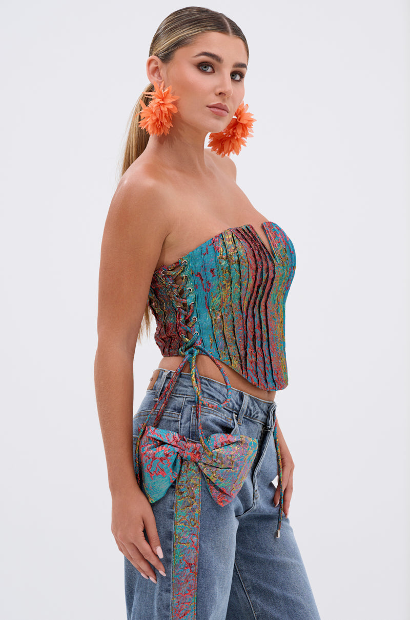 A woman with long hair in a ponytail wears large orange flower earrings, blue jeans with a matching bow, and the PRETTY GIRL PROBLEMS CORSET—a colorful strapless metallic party top—while standing against a plain white background.