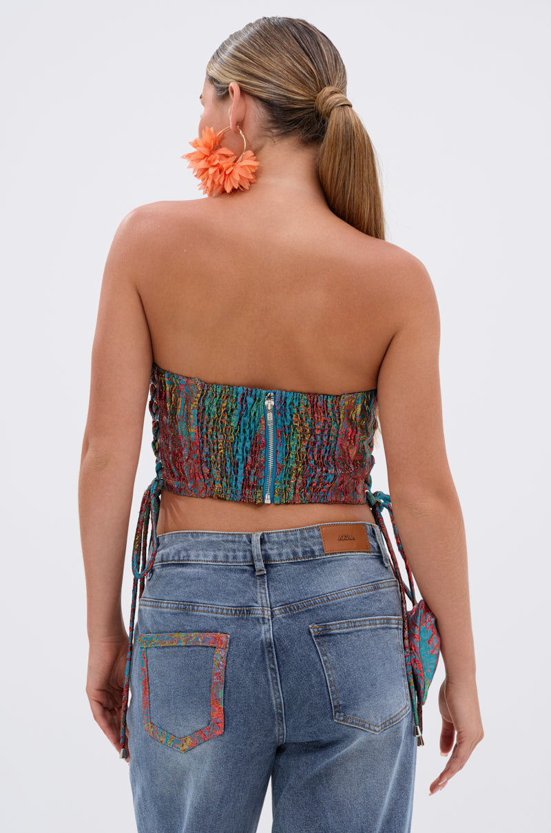 A woman with long, straight hair in a low ponytail is shown from the back wearing the PRETTY GIRL PROBLEMS CORSET, high-waisted blue jeans, large orange flower earrings, and holding a small matching bag.