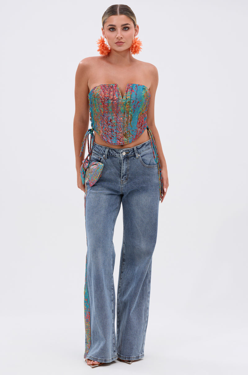 A woman wears large orange flower earrings, the PRETTY GIRL PROBLEMS CORSET— a strapless sweetheart crop top with a pointed hem— and high-waisted blue jeans featuring a colorful patch and wide legs, standing against a white background.