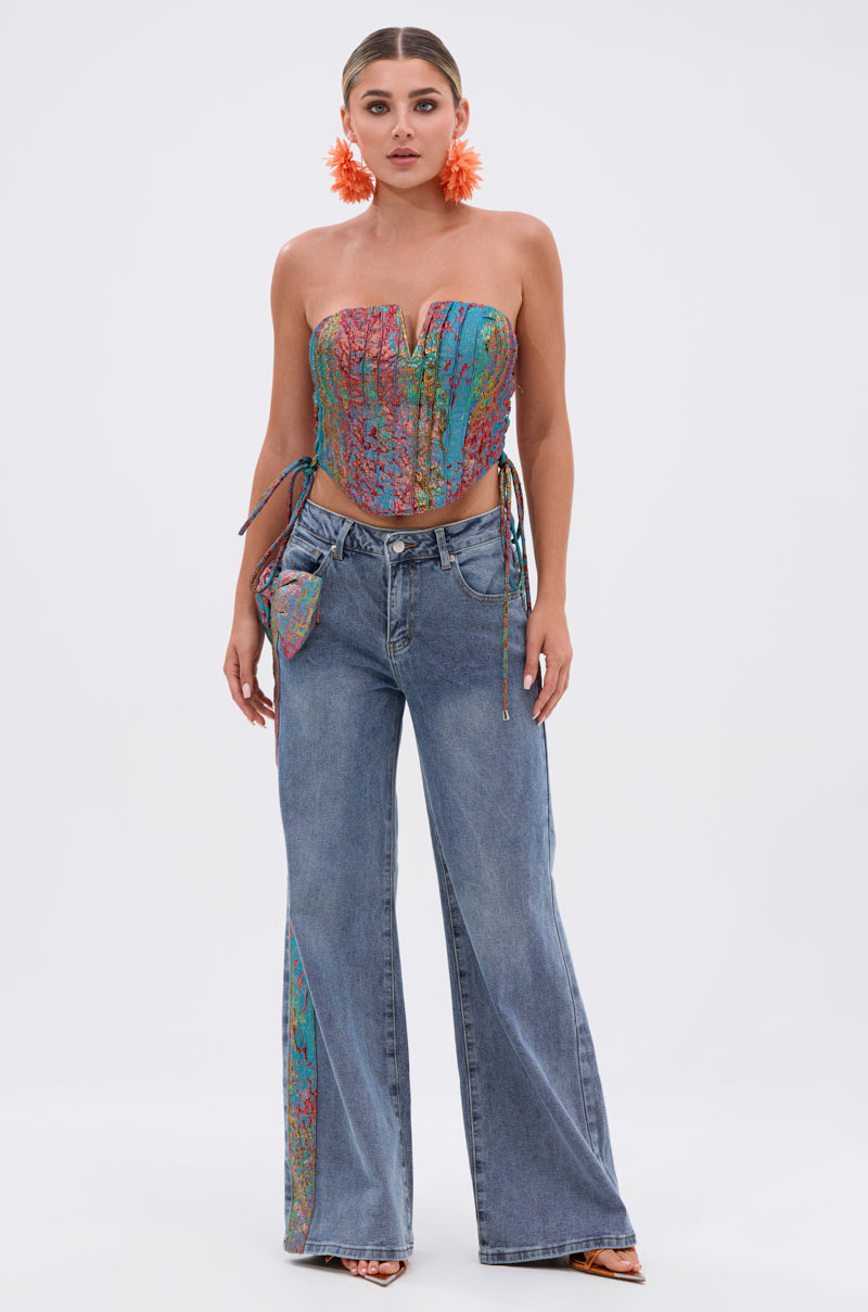 A woman wears bold orange floral earrings, the PRETTY GIRL PROBLEMS CORSET with a colorful patterned strapless sweetheart neckline, and high-waisted wide-leg jeans featuring matching details and an attached pouch, posed against a plain white background.