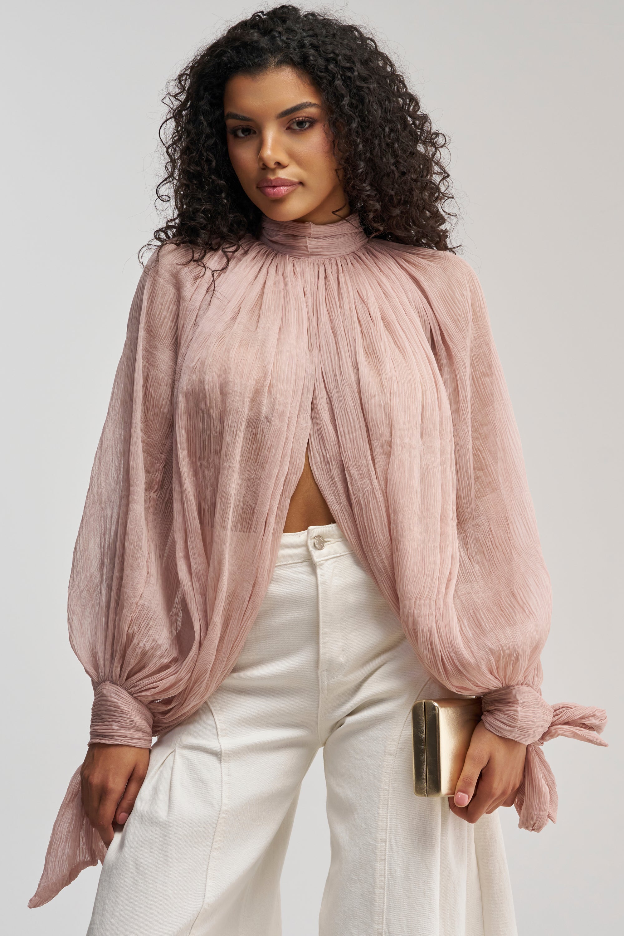 Wearing the SOFT SEDUCTION CHIFFON BLOUSE in delicate pink—with sheer sleeves, pleated details, and tied cuffs—a woman with curly hair pairs it with white high-waisted pants and carries a gold clutch.