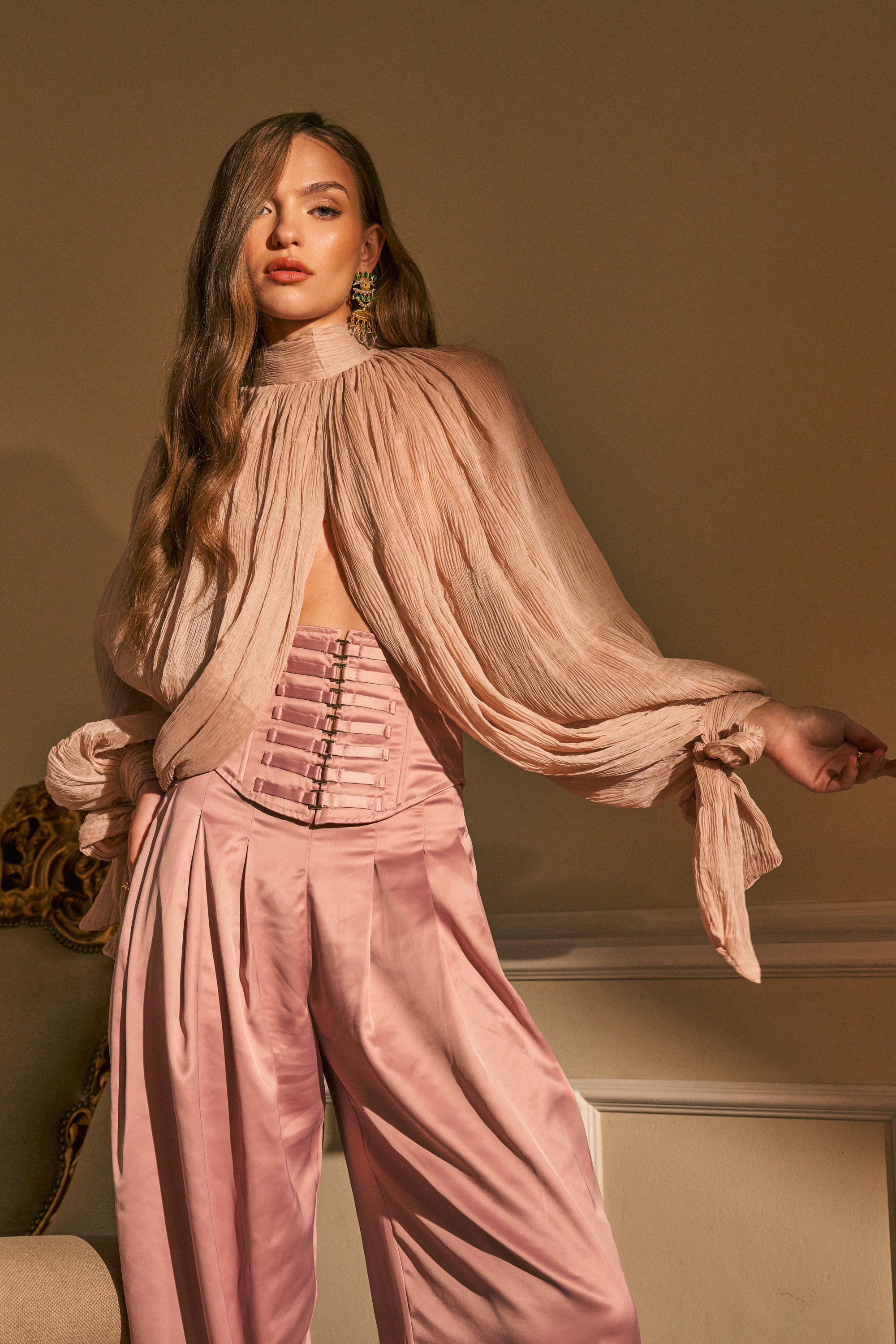 A woman with long wavy hair wears the SOFT SEDUCTION CHIFFON BLOUSE—a light pink mock neck top with billowy sleeves—paired with matching high-waisted pleated pants, posing confidently indoors in soft lighting.