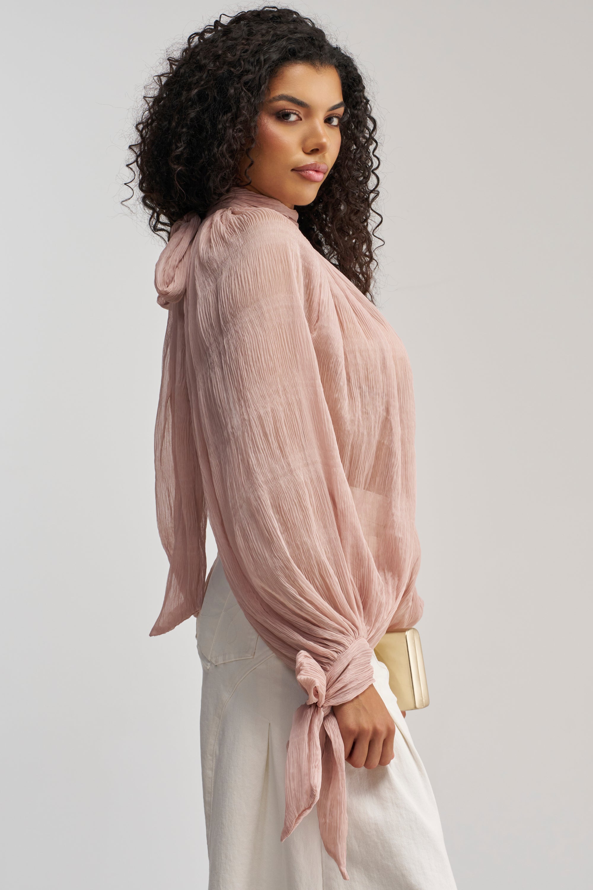 A woman with curly hair stands in profile against a plain background, wearing the SOFT SEDUCTION CHIFFON BLOUSE in mauve with tied sleeves, paired with white pants and holding a light-colored clutch.