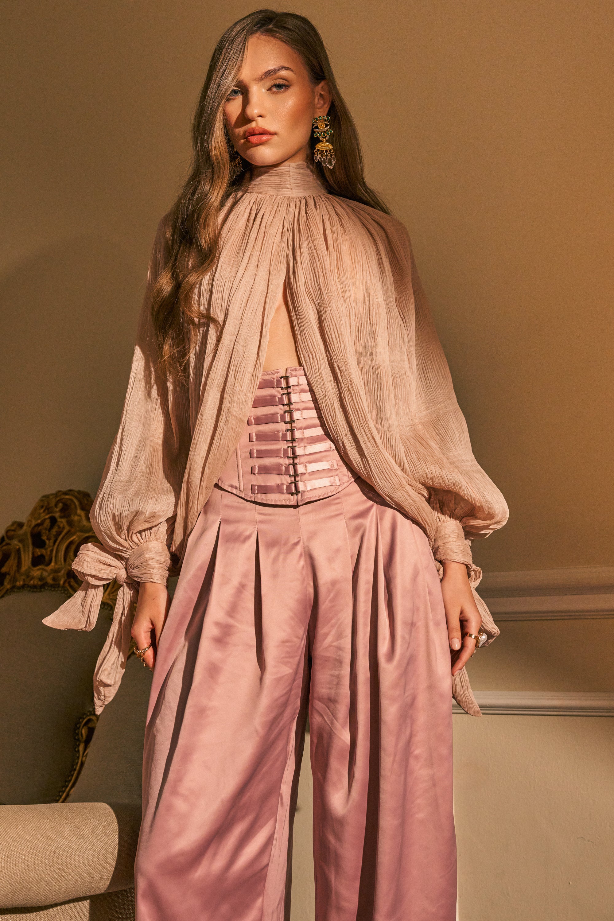 A woman wearing the SOFT SEDUCTION CHIFFON BLOUSE in mauve, featuring a high neck and tied sleeves, paired with wide-legged satin pants and statement earrings, stands indoors near a chair.