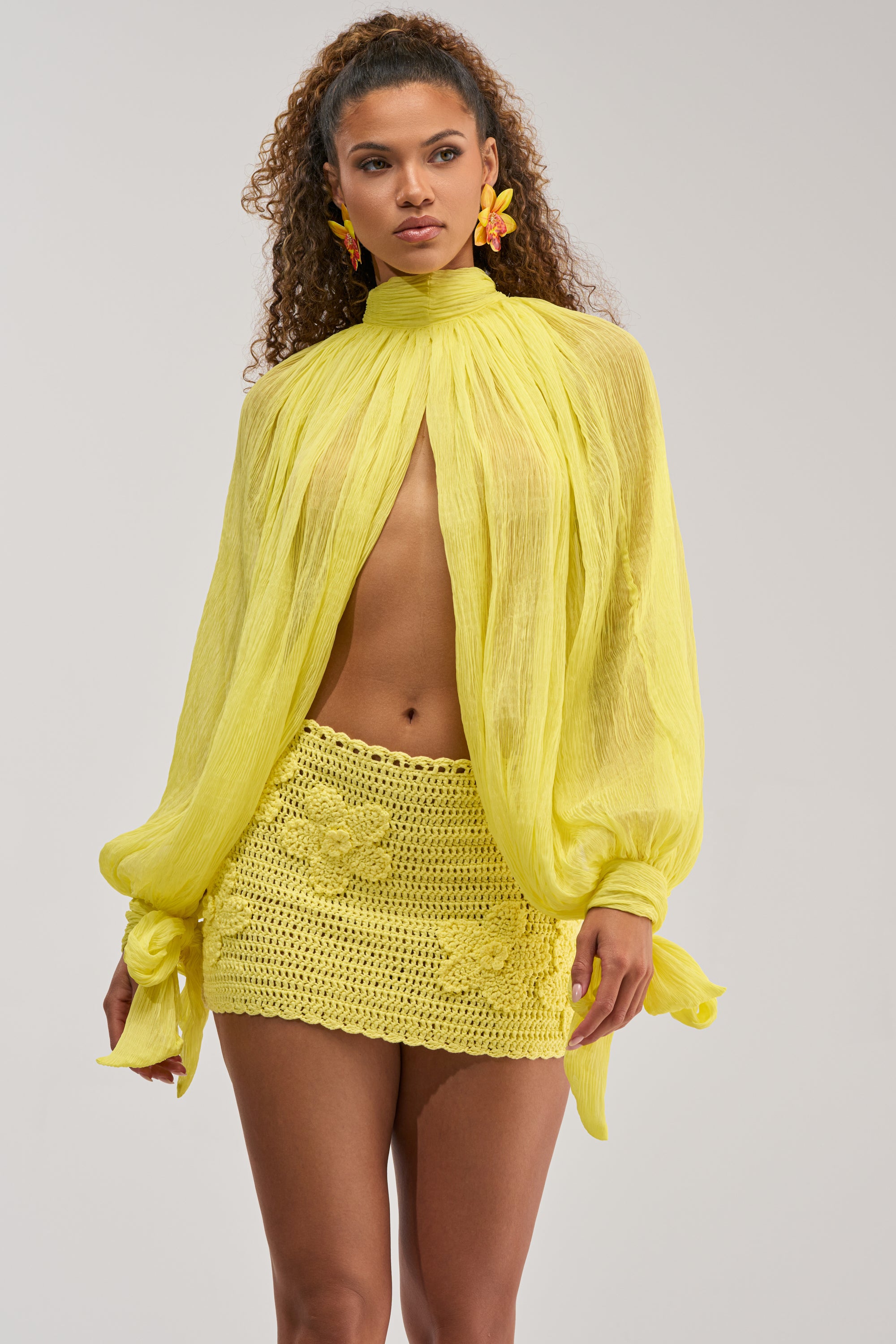 A woman poses in the HELLO SUNSHINE CHIFFON BLOUSE—a sheer, long-sleeve yellow top with tied cuffs—styled with a crocheted floral mini skirt and flower-shaped earrings, her curly hair standing out against a plain backdrop.