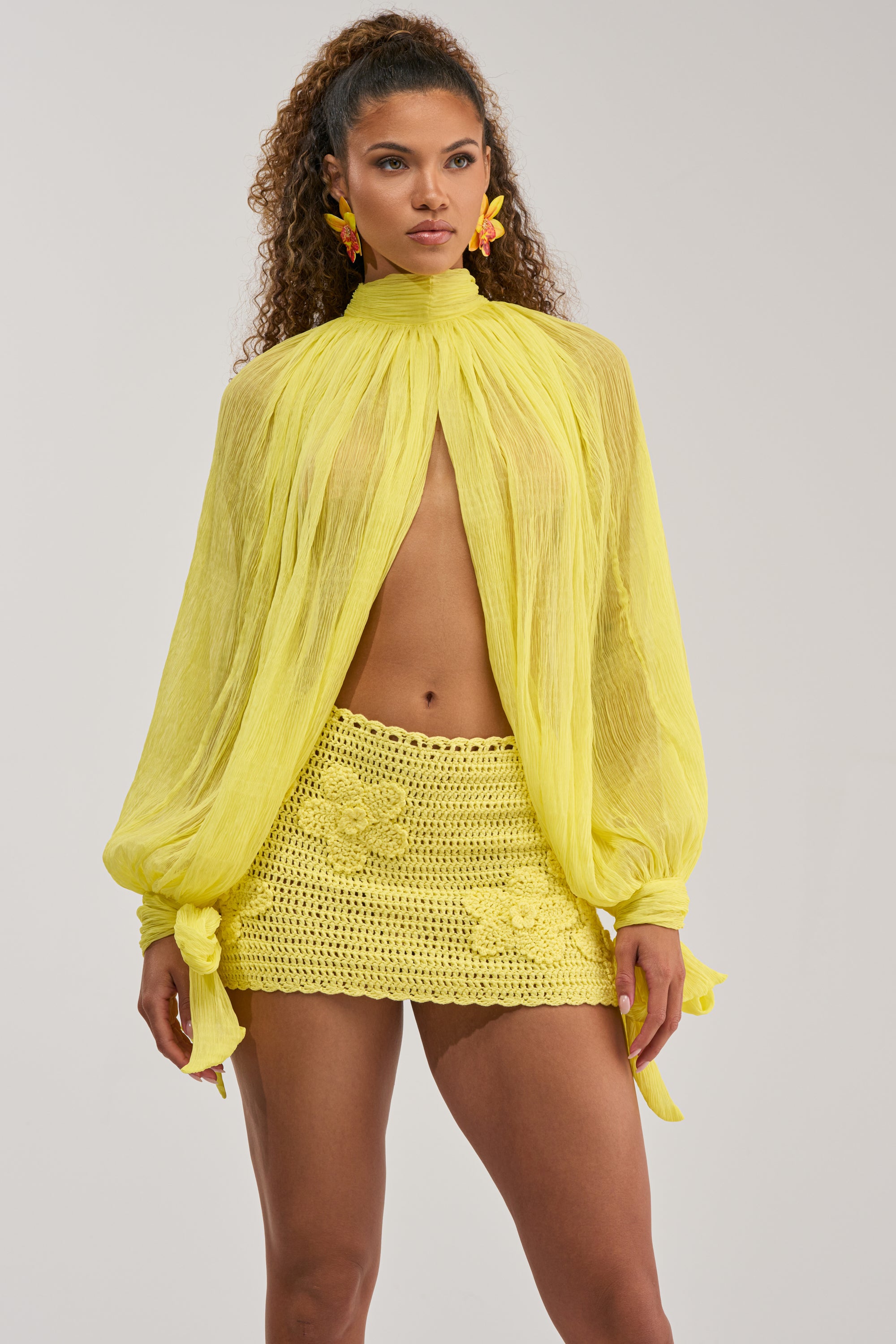 A woman wears the HELLO SUNSHINE CHIFFON BLOUSE—a sheer, long-sleeved yellow top with a high neck and bow details—paired with a yellow crocheted mini skirt adorned with floral patterns. With her curly hair, statement earrings, and confident expression, she’s ready to make an entrance in this summer statement piece.