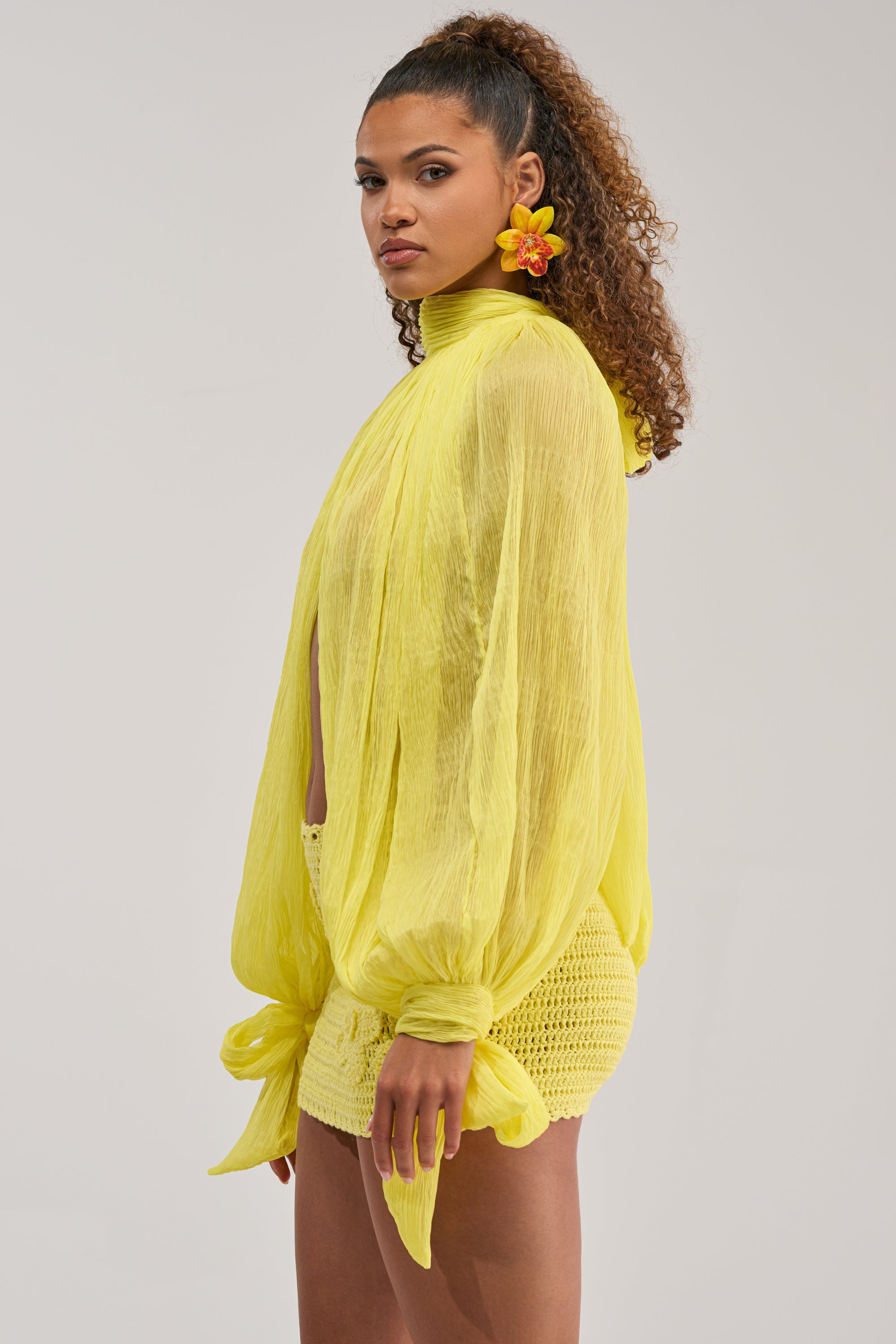 A woman with curly hair and a yellow flower wears the HELLO SUNSHINE CHIFFON BLOUSE with matching crochet shorts, standing sideways against a plain light background—an eye-catching statement summer look.