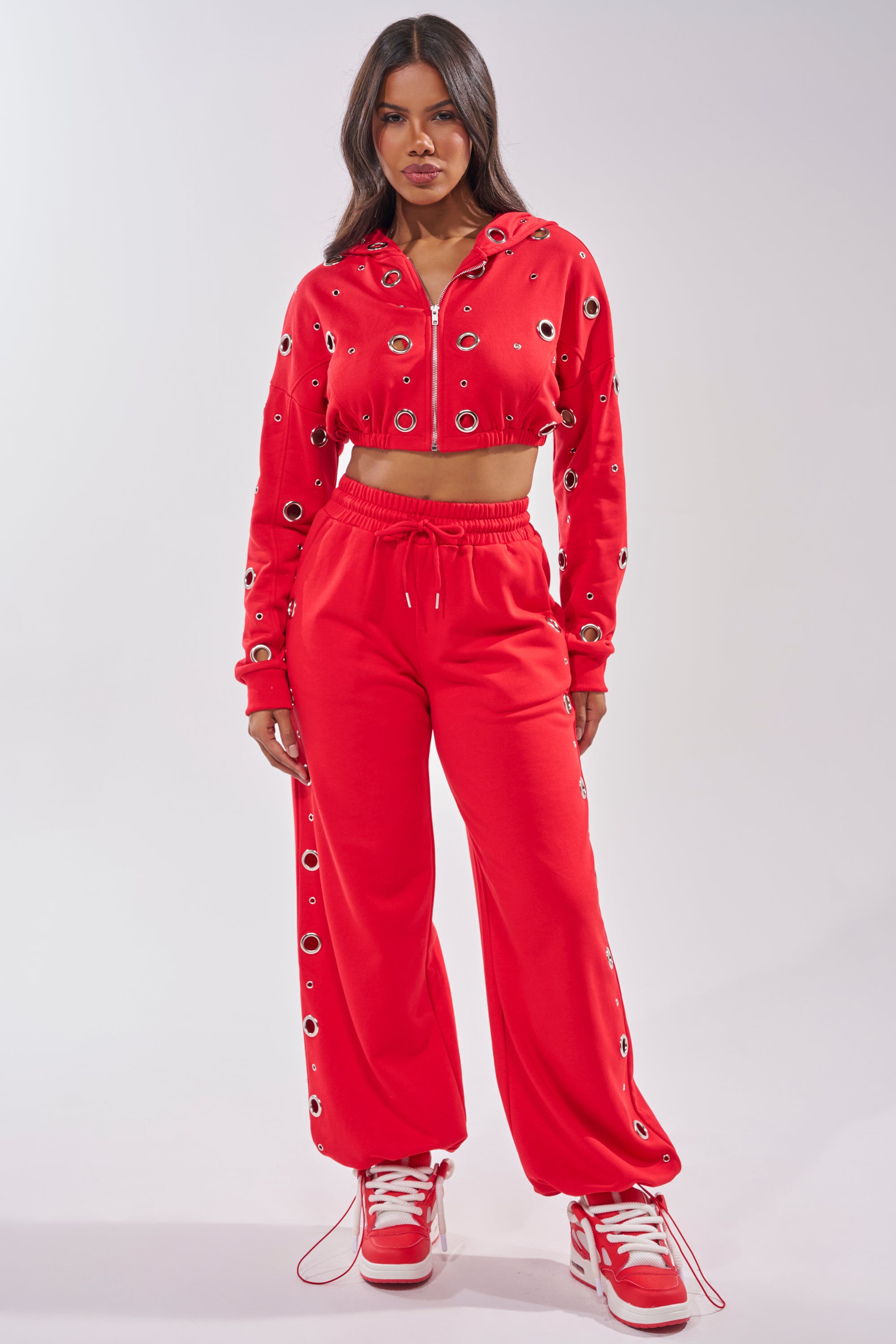 A woman faces forward in a bright red cropped hoodie and COMFY COZY SWEATPANT with eyelet details, paired with red and white sneakers. The sporty, casual look features the hoodie revealing her midriff. 10