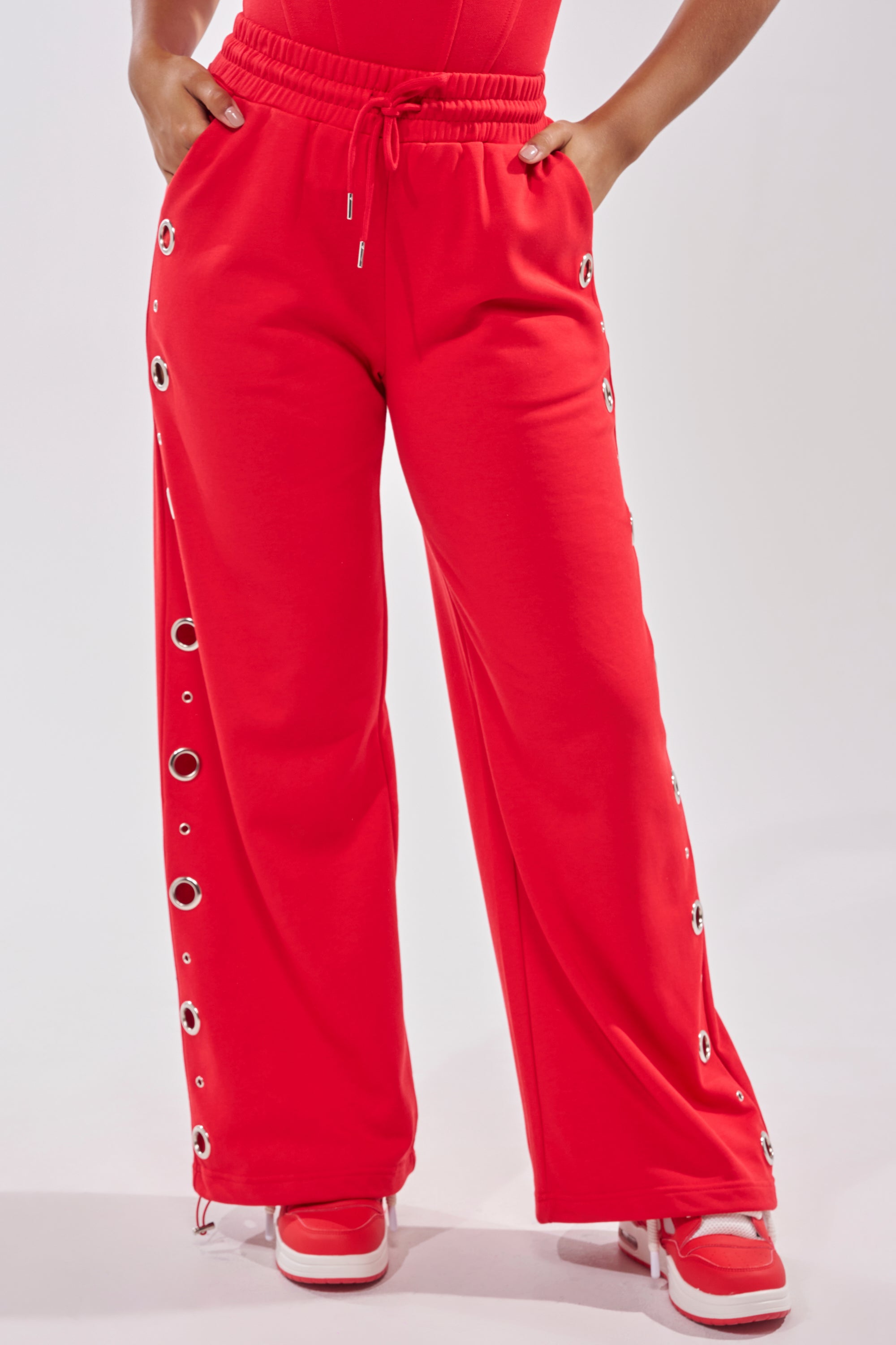 A person wears the COMFY COZY SWEATPANT in bright red, featuring wide legs and silver grommet details along the sides, paired with matching red sneakers. The image is cropped from the waist down. 2