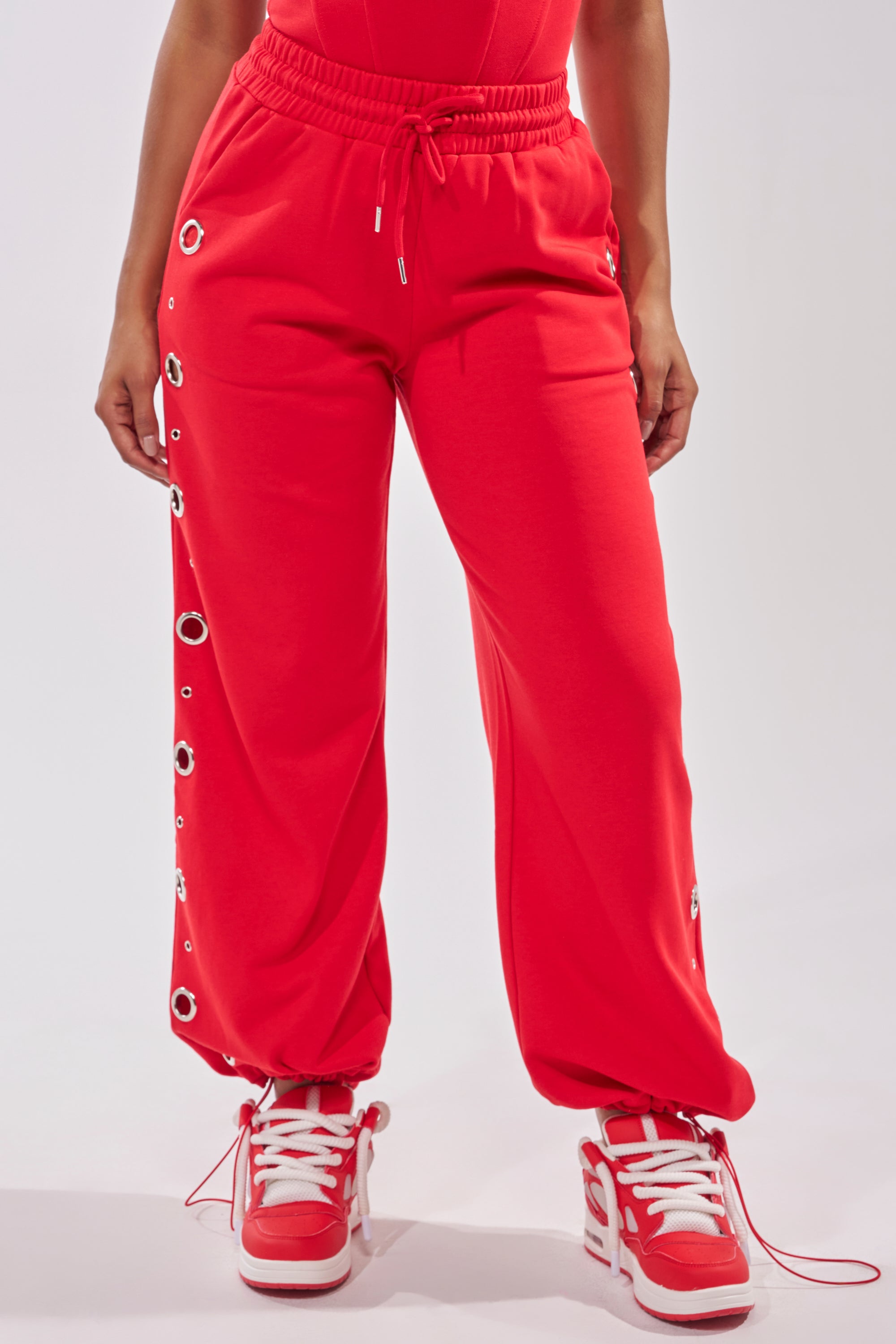 Someone is wearing bright red COMFY COZY SWEATPANT featuring silver grommet details along one leg, paired with red and white sneakers against a plain, light-colored background. 3