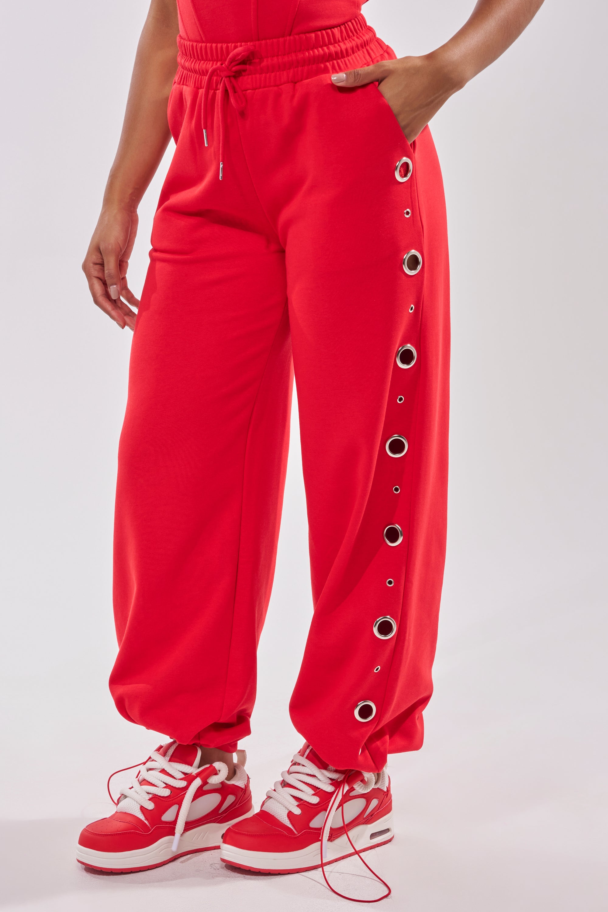 Someone stands against a plain white background wearing COMFY COZY SWEATPANT in bright red cotton with silver details on one leg, paired with red and white sneakers. 4