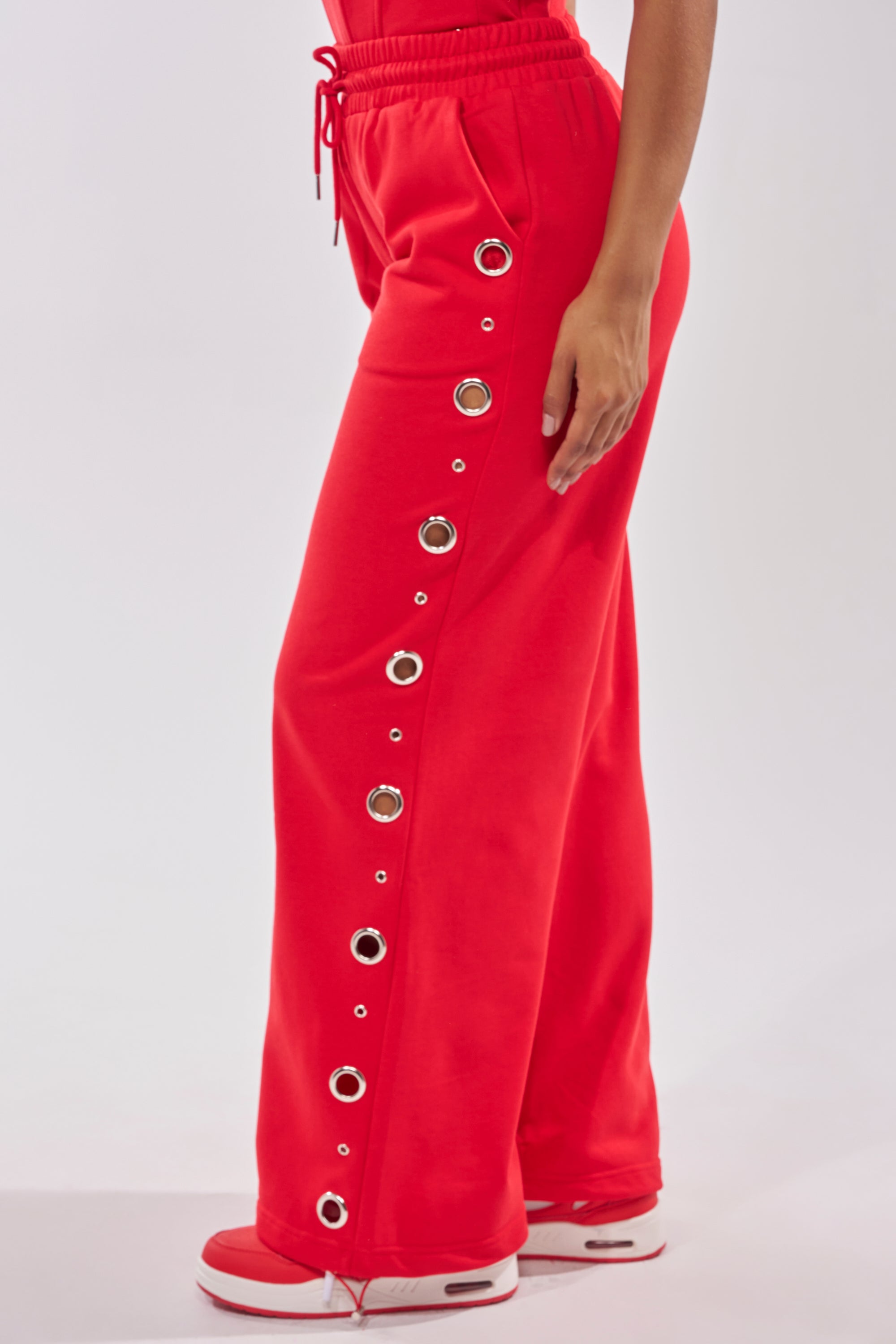 Someone stands against a plain light background wearing the COMFY COZY SWEATPANT in bright red with large silver grommets down the side seam, paired with red-and-white sneakers. 5