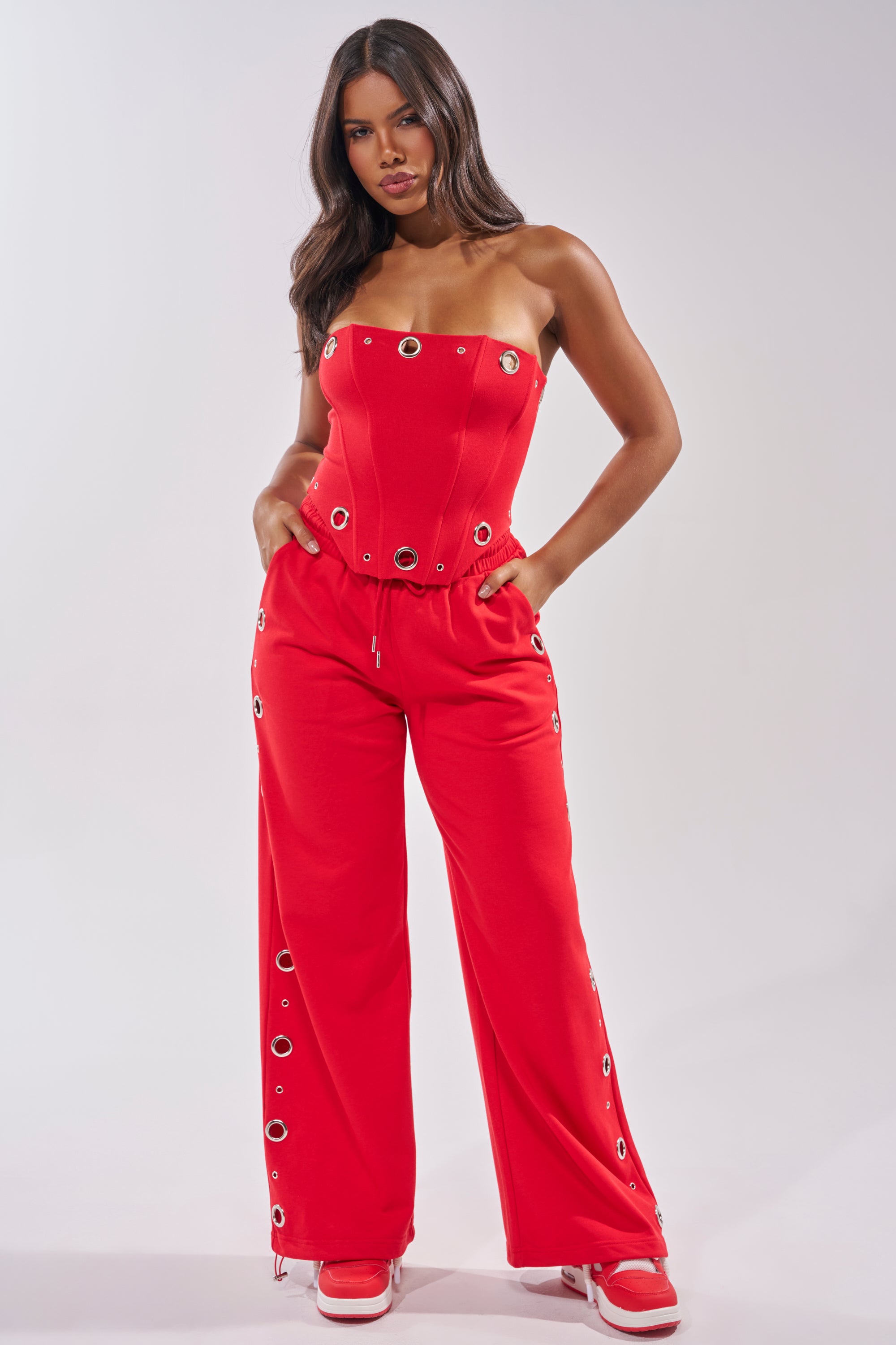 A woman models the COMFY COZY CORSET, a strapless grommet corset top with silver accents, styled with matching red wide-leg grommet pants and red and white sneakers, set against a plain light background. 6