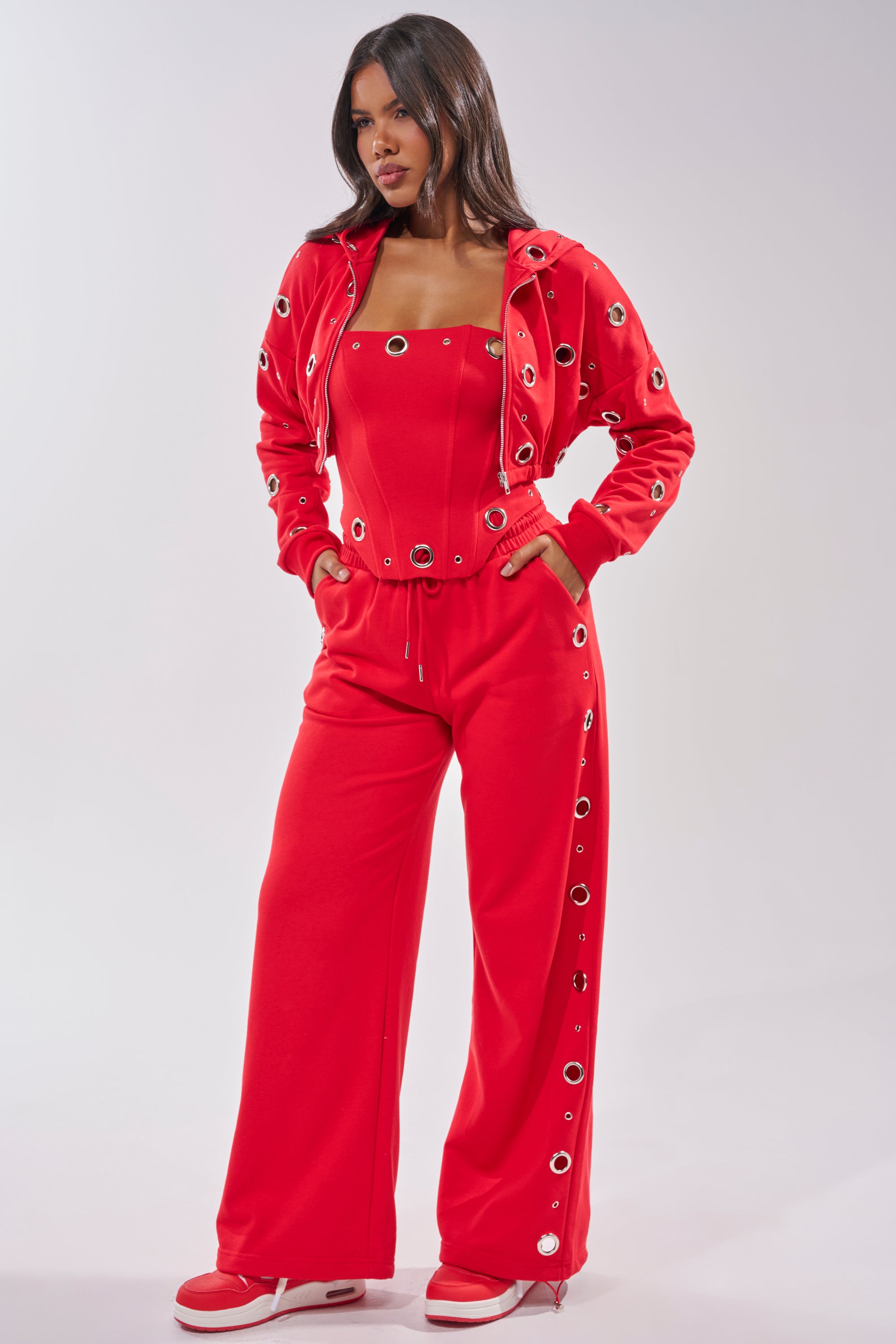 A woman exudes confidence in the COMFY COZY CORSET, a bright red strapless top paired with wide-leg pants featuring large metallic grommets. Her hands rest in her pockets, and she completes the bold look with red and white sneakers. 7