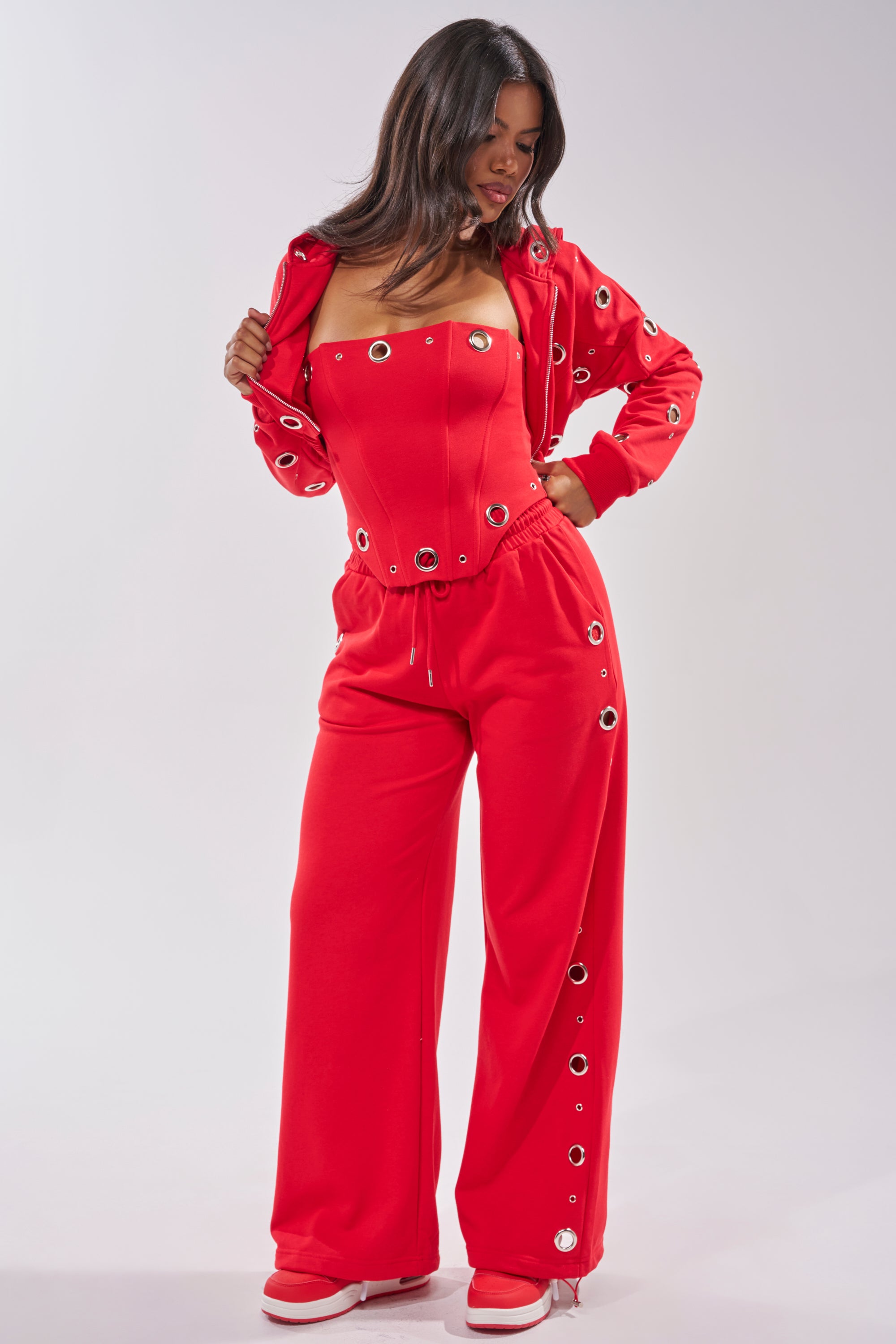 Woman wearing the COMFY COZY CORSET in vibrant red cotton, styled with wide-leg pants and a matching jacket embellished with metallic grommets. She stands looking down, holding her jacket in one hand. 8