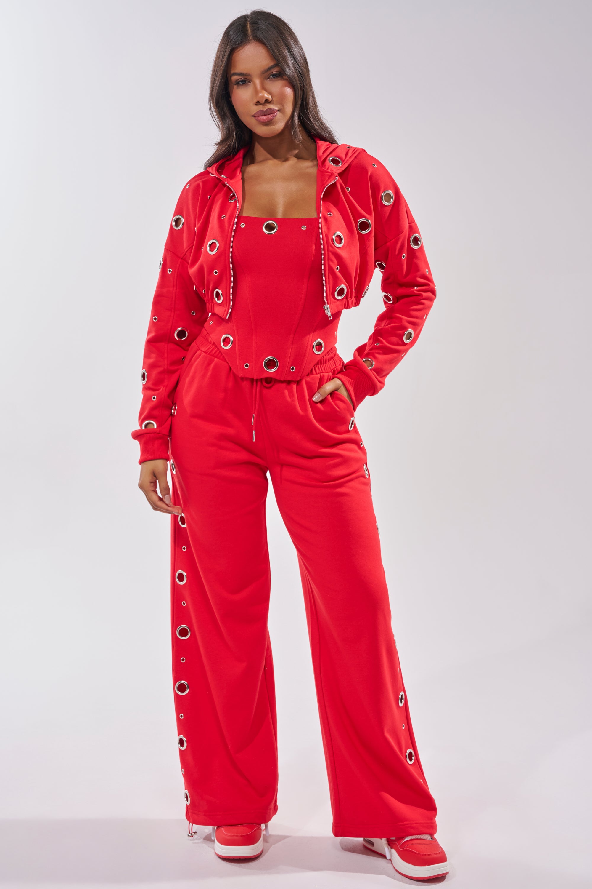 A woman stands confidently in a bright red matching set featuring the COMFY COZY CROPPED HOODIE with bold grommet details, a tank top, wide-leg pants, and red and white sneakers against a plain white background. 10