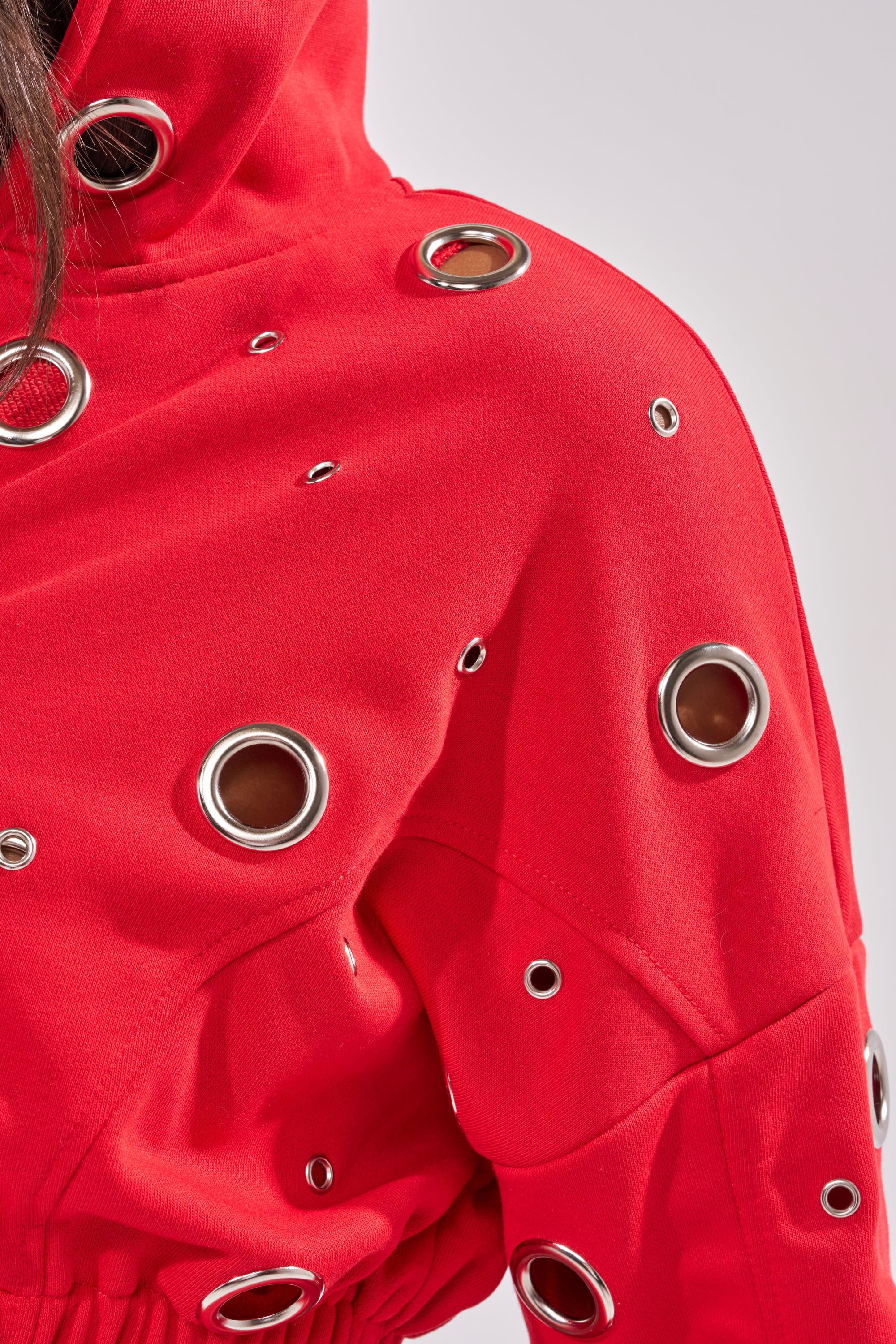 Close-up of the COMFY COZY CROPPED HOODIE in red, featuring bold silver grommet accents throughout its cotton fabric; partial view of the hood and sleeve. 13