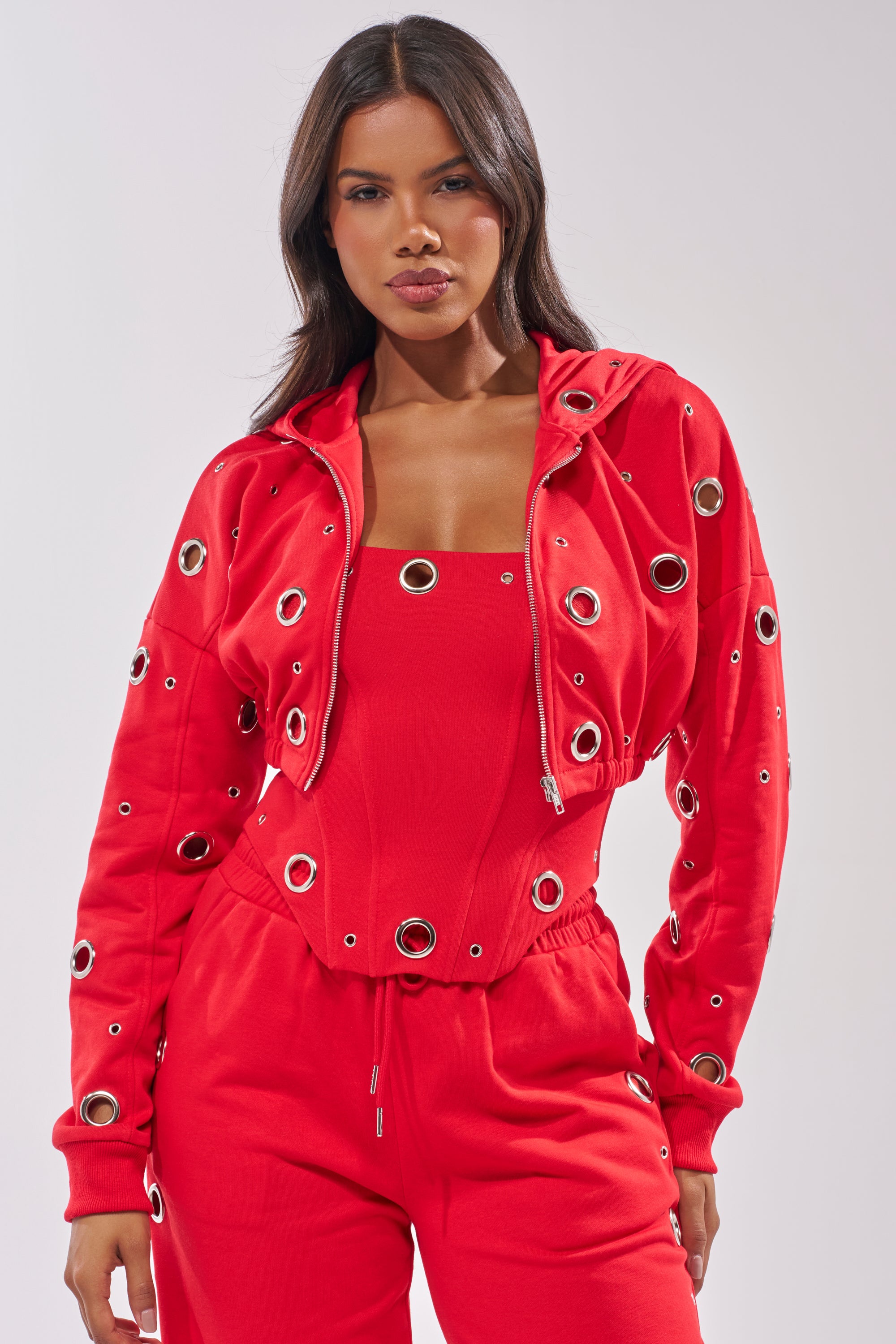 A woman wears the COMFY COZY CROPPED HOODIE and matching pants in red, both with bold grommet detailing. She stands facing forward with a neutral expression against a plain light background. 2