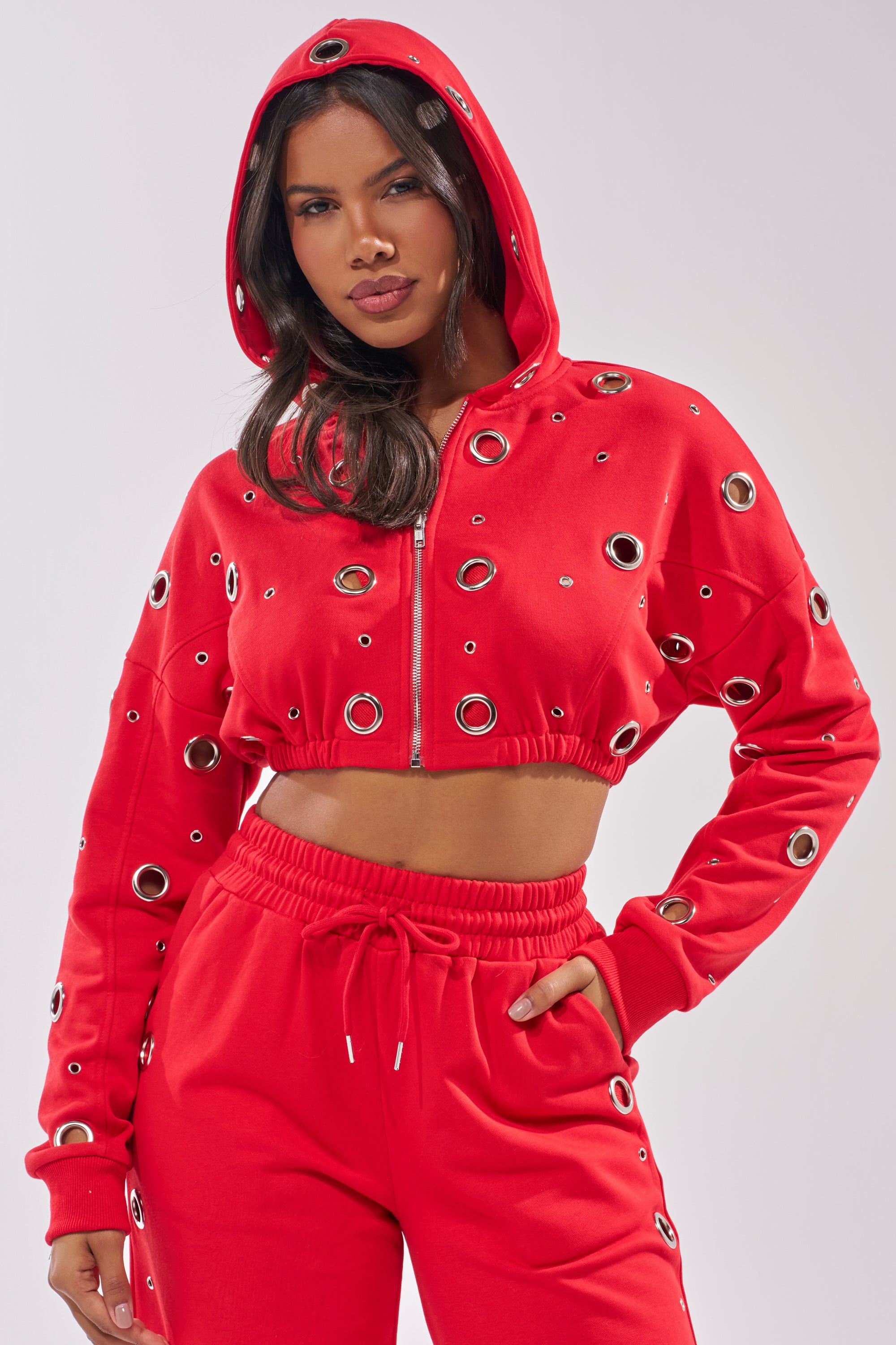 A woman poses with one hand on her hip, wearing the COMFY COZY CROPPED HOODIE and matching sweatpants with eye-catching silver grommet details. 5