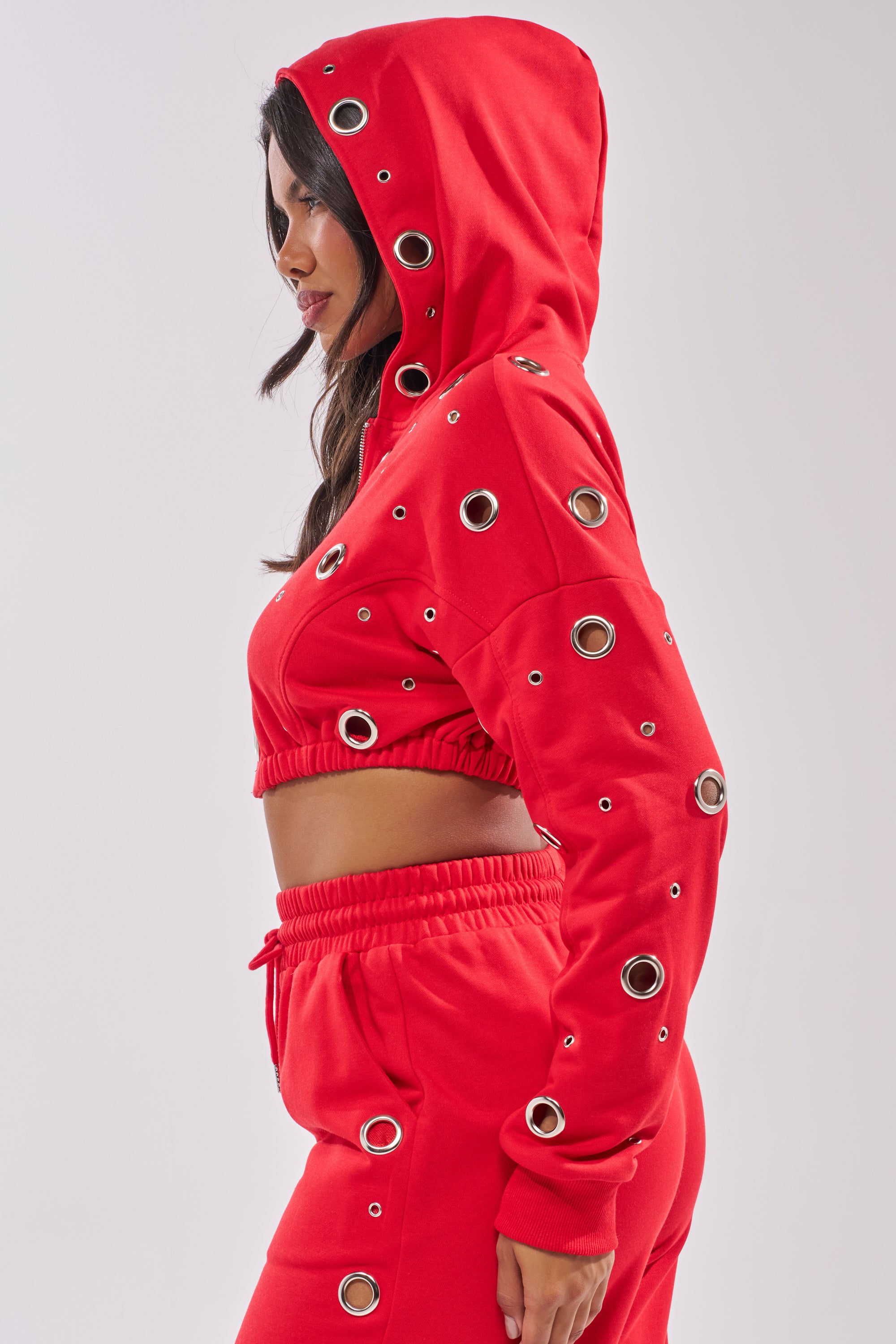 A woman faces sideways wearing the COMFY COZY CROPPED HOODIE in red, featuring cotton fabric, grommet details, a drawstring, and matching sweatpants with an elastic waistband. 6