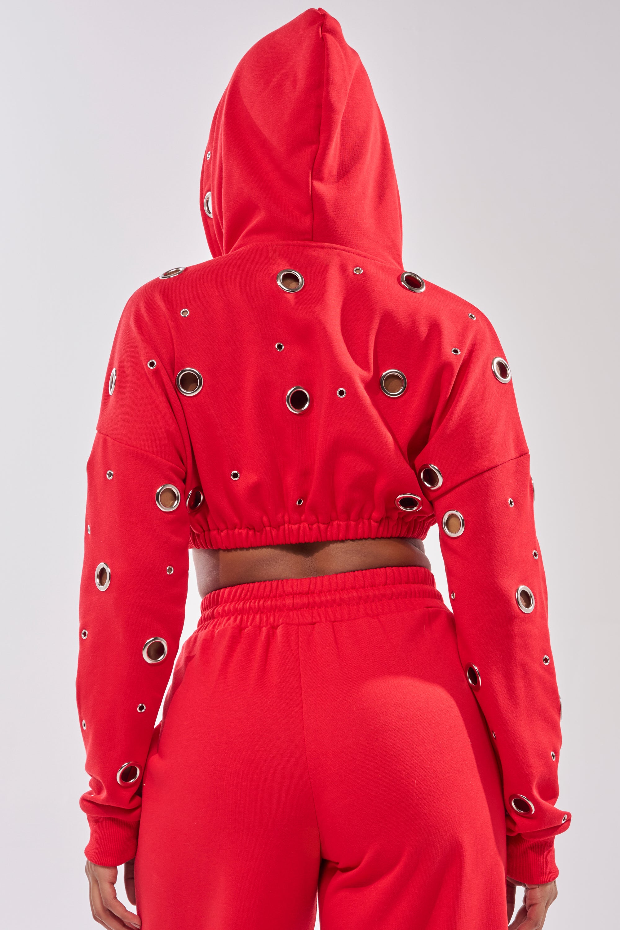 A person shown from the back wears the COMFY COZY CROPPED HOODIE in red, featuring silver grommet details, an elastic waistband, and matching red pants for a bold, modern look. 7