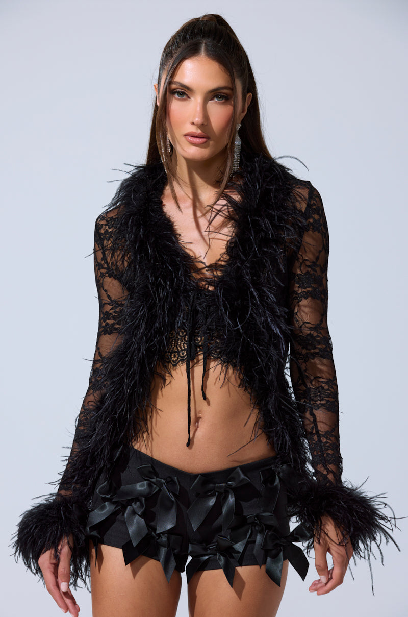 A woman models the SEXY LACE BLOUSE with feather trim and black shorts featuring satin bows, standing against a plain light background. 3