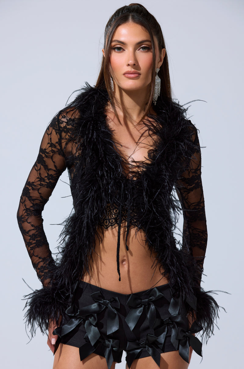 A woman wears the SEXY LACE BLOUSE, made of sheer black lace with feather trim and long sleeves, paired with black shorts featuring satin bows. She has straight hair, statement earrings, and stands before a plain light background. 0