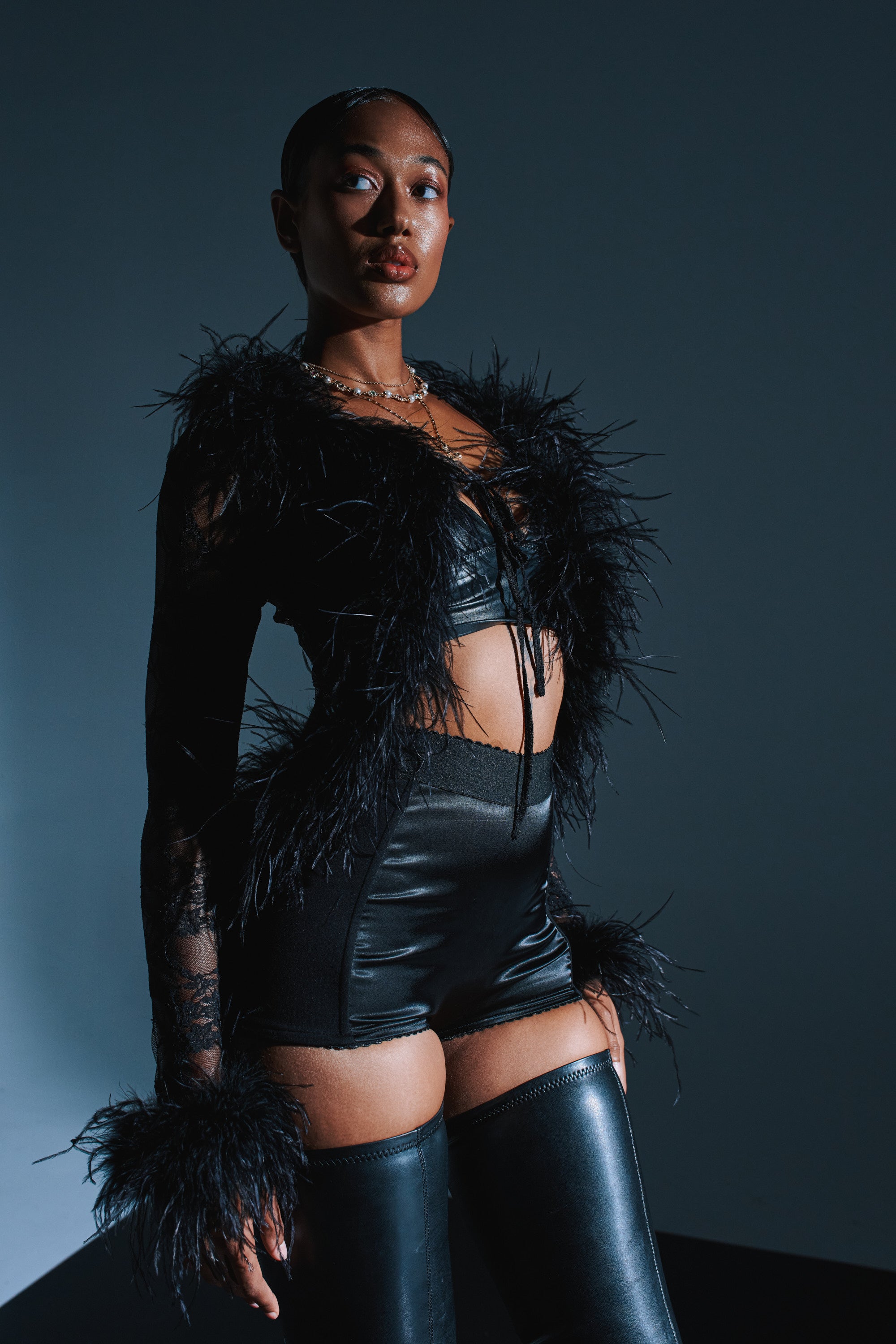 A woman exudes confidence in the SEXY LACE BLOUSE—a sheer black lace top with feather trim—paired with black leather shorts and thigh-high boots, posing against a dark, minimalist backdrop under dramatic lighting. 2