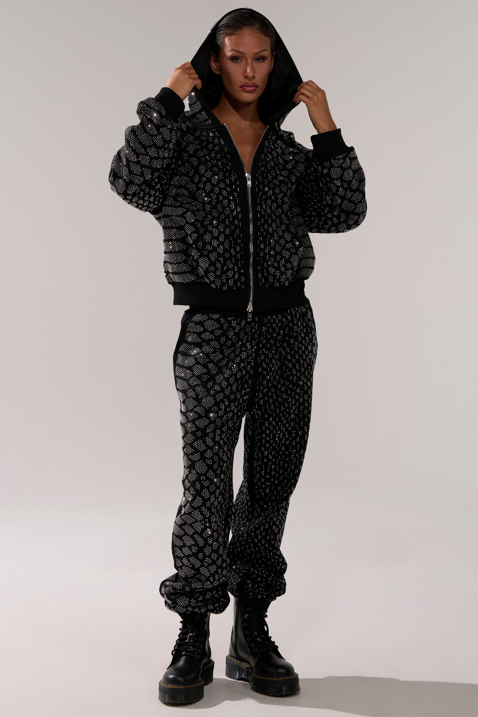 A person stands confidently in a black hooded bomber jacket, SNEAKY LITTLE SNAKE WIDE LEG JOGGER pants with a sparkling geometric pattern, and black boots.