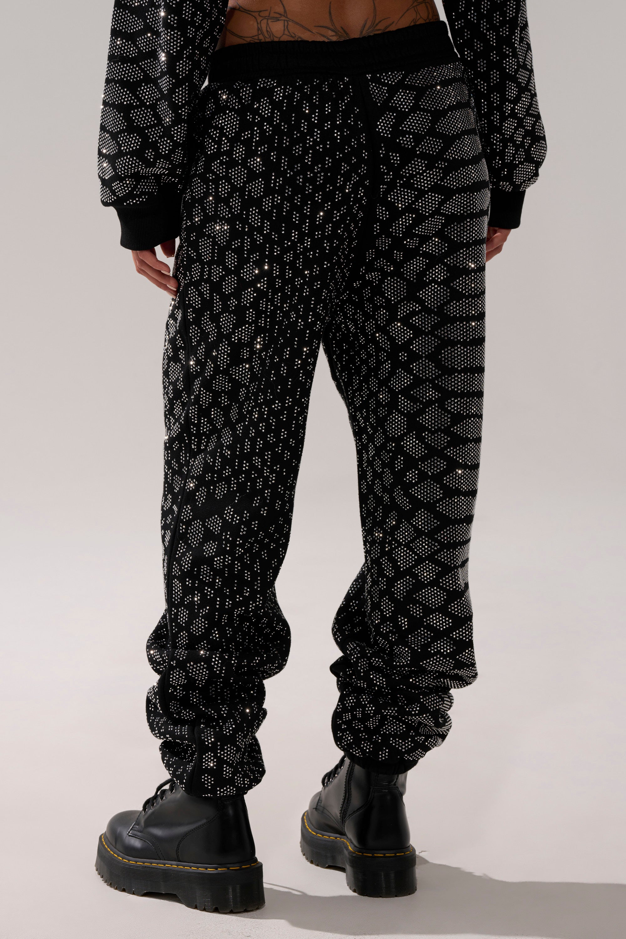 A person faces away wearing the SNEAKY LITTLE SNAKE WIDE LEG JOGGER—black joggers with a geometric silver pattern—styled with black boots and a matching top, set against a plain, neutral background.