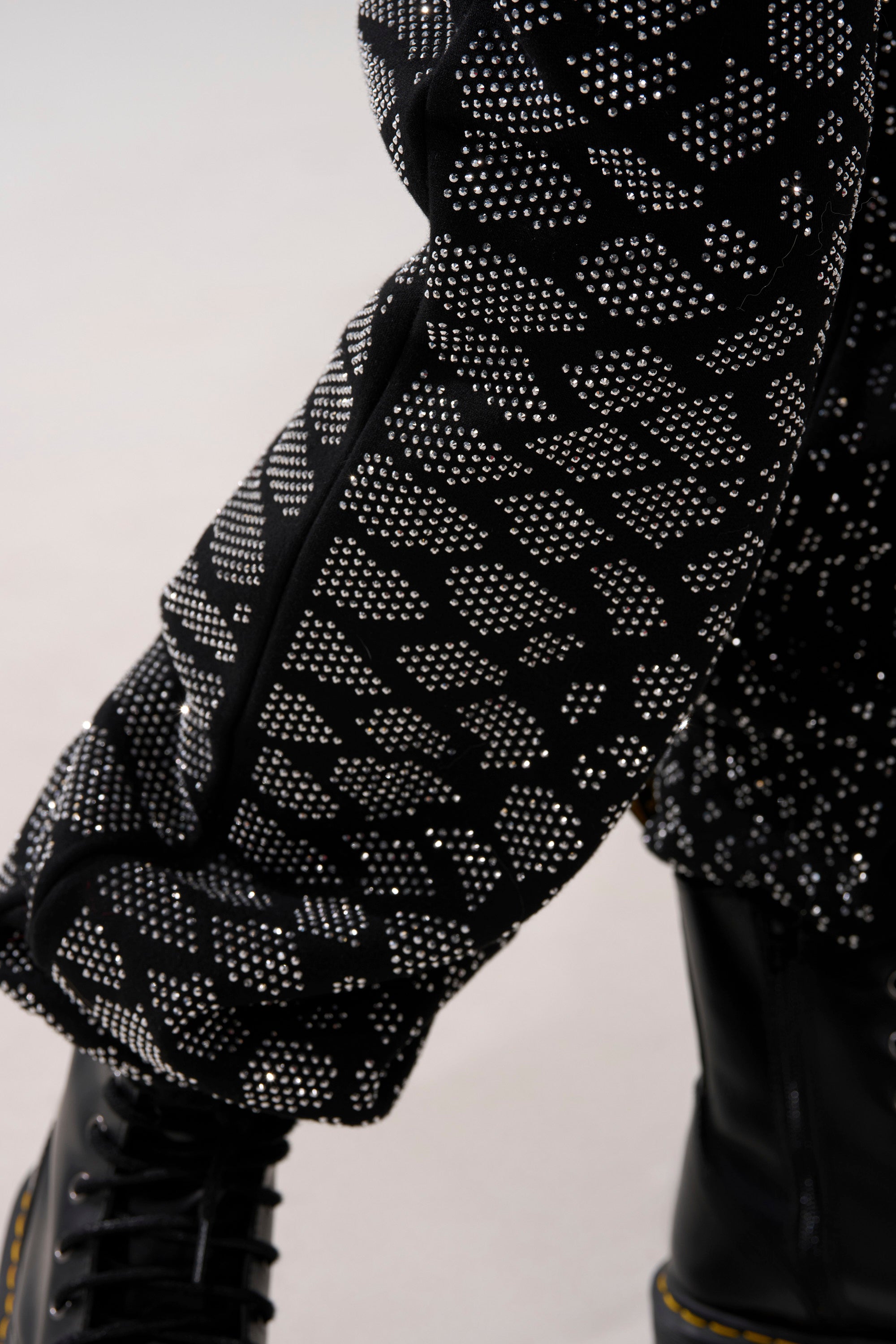 A close-up of a leg wearing the SNEAKY LITTLE SNAKE WIDE LEG JOGGER—black jogger pants with a geometric white dot pattern—paired with black leather boots featuring yellow stitching.
