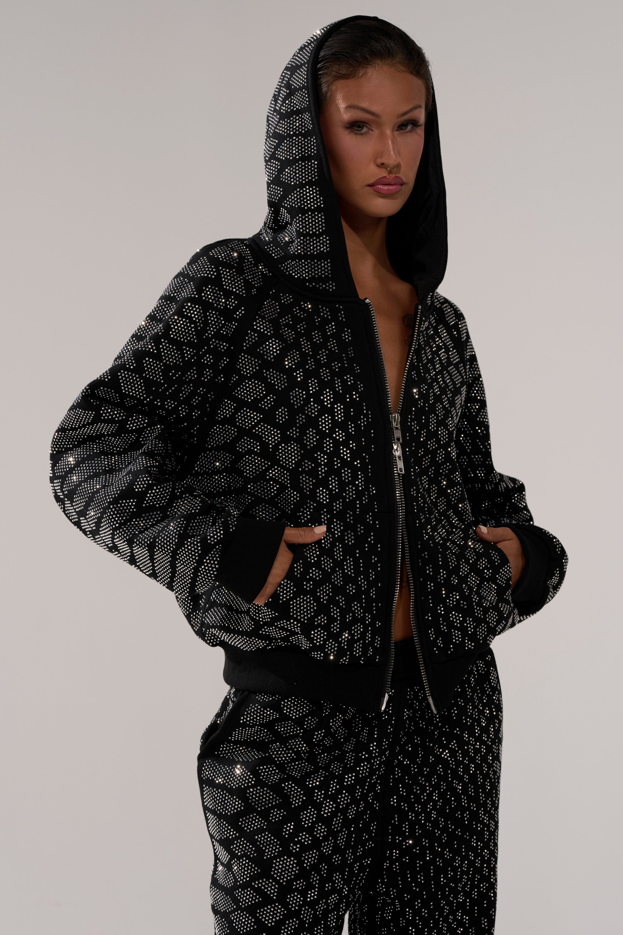 A woman stands facing forward with her hands in her pockets, wearing the SNEAKY LITTLE SNAKE ZIP UP SWEATSHIRT and matching pants featuring a silver geometric pattern, posed against a plain light background.