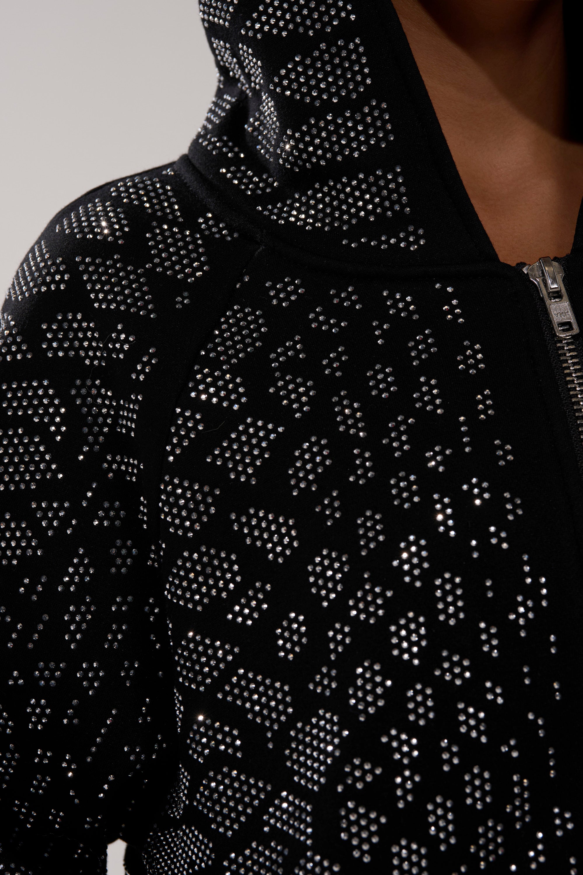 Close-up of the SNEAKY LITTLE SNAKE ZIP UP SWEATSHIRT, a black zip-up hoodie with a hood, detailed with shiny silver studs in a geometric pattern. Part of a person’s face and neck appear in the upper right corner.