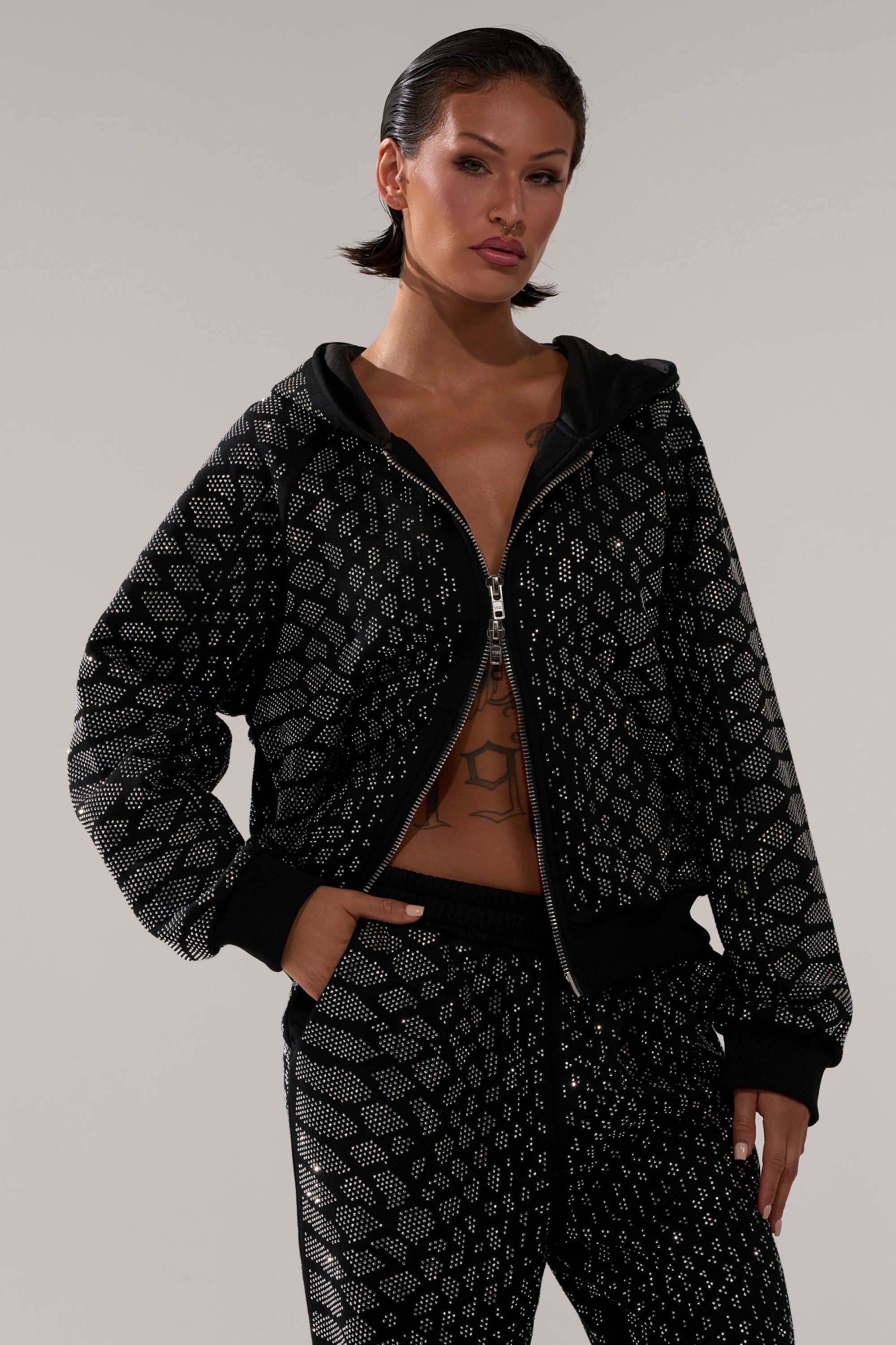 A woman with slicked-back hair confidently models the SNEAKY LITTLE SNAKE ZIP UP SWEATSHIRT and matching geometric silver pants, one hand in her pocket, highlighting the cozy hoodie against a plain background.